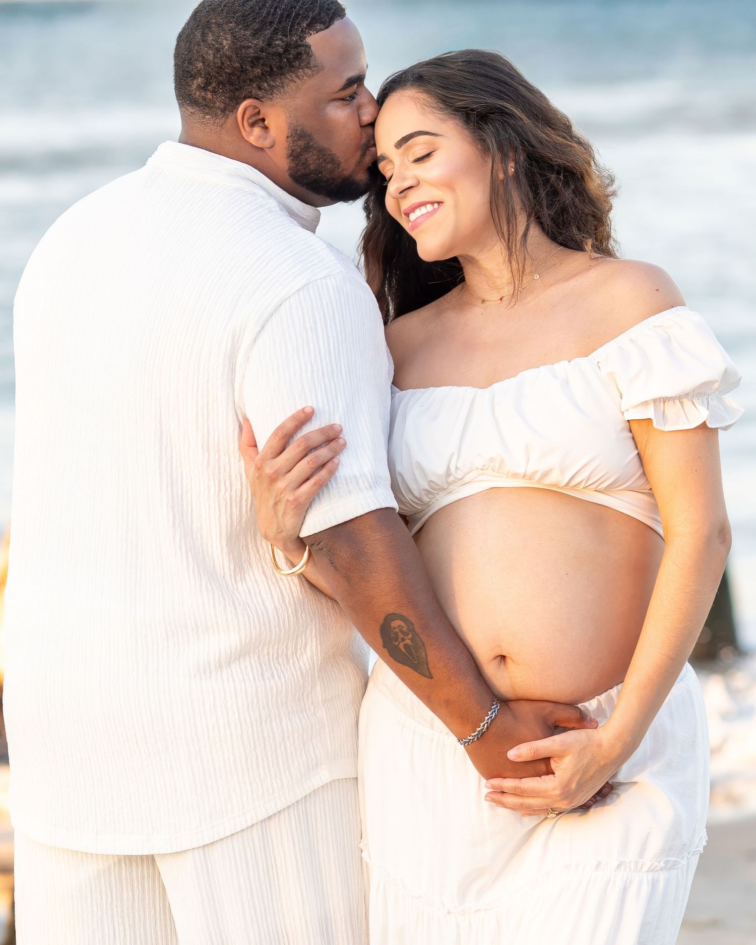 Romantic maternity photo of couple in white outfits embracing on beach at sunset.