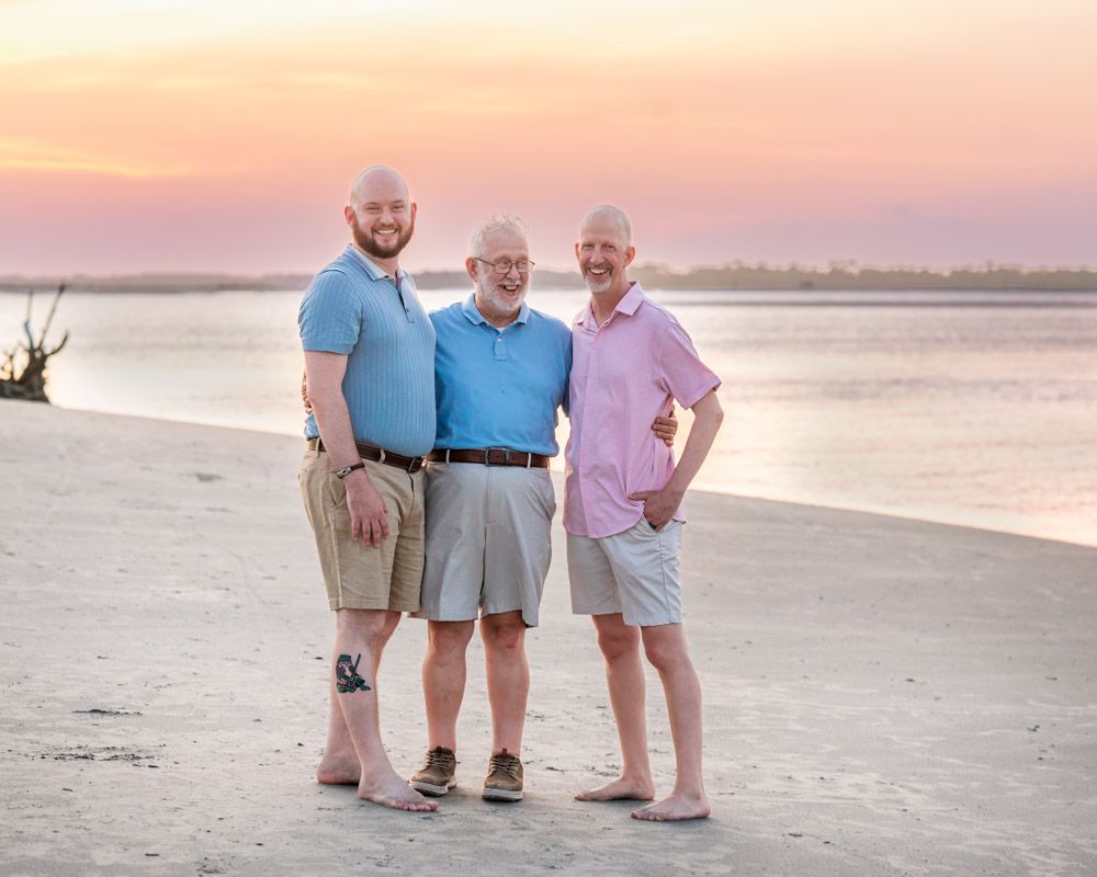 Three men smiling together on a sandy beach at sunset with a colorful pink and orange sky backdrop.