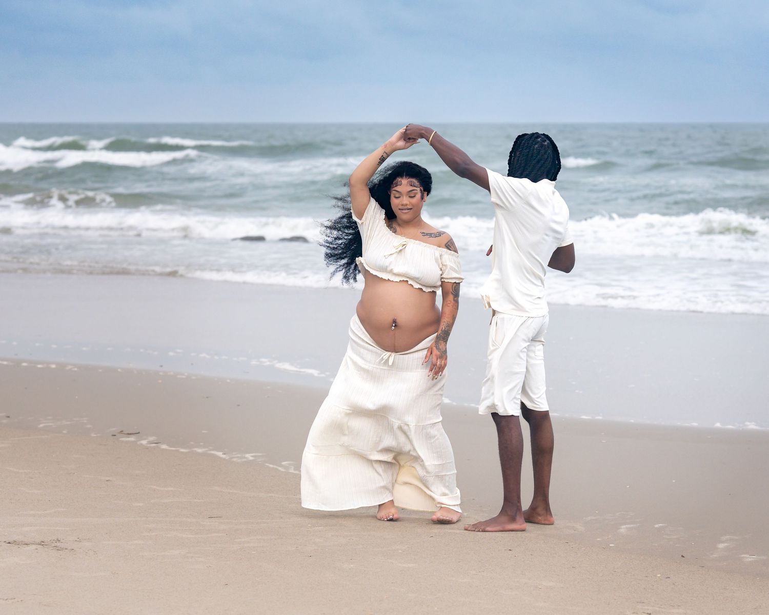 Pregnant woman in white dress dances with partner on beach, waves in background.