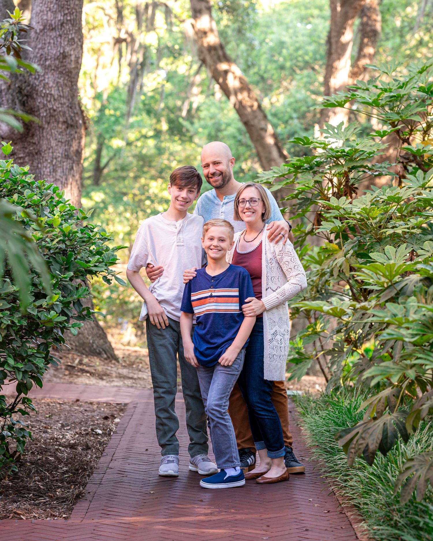 Family of four smiling together on a wooden path surrounded by lush green trees and shrubs in a park setting.
