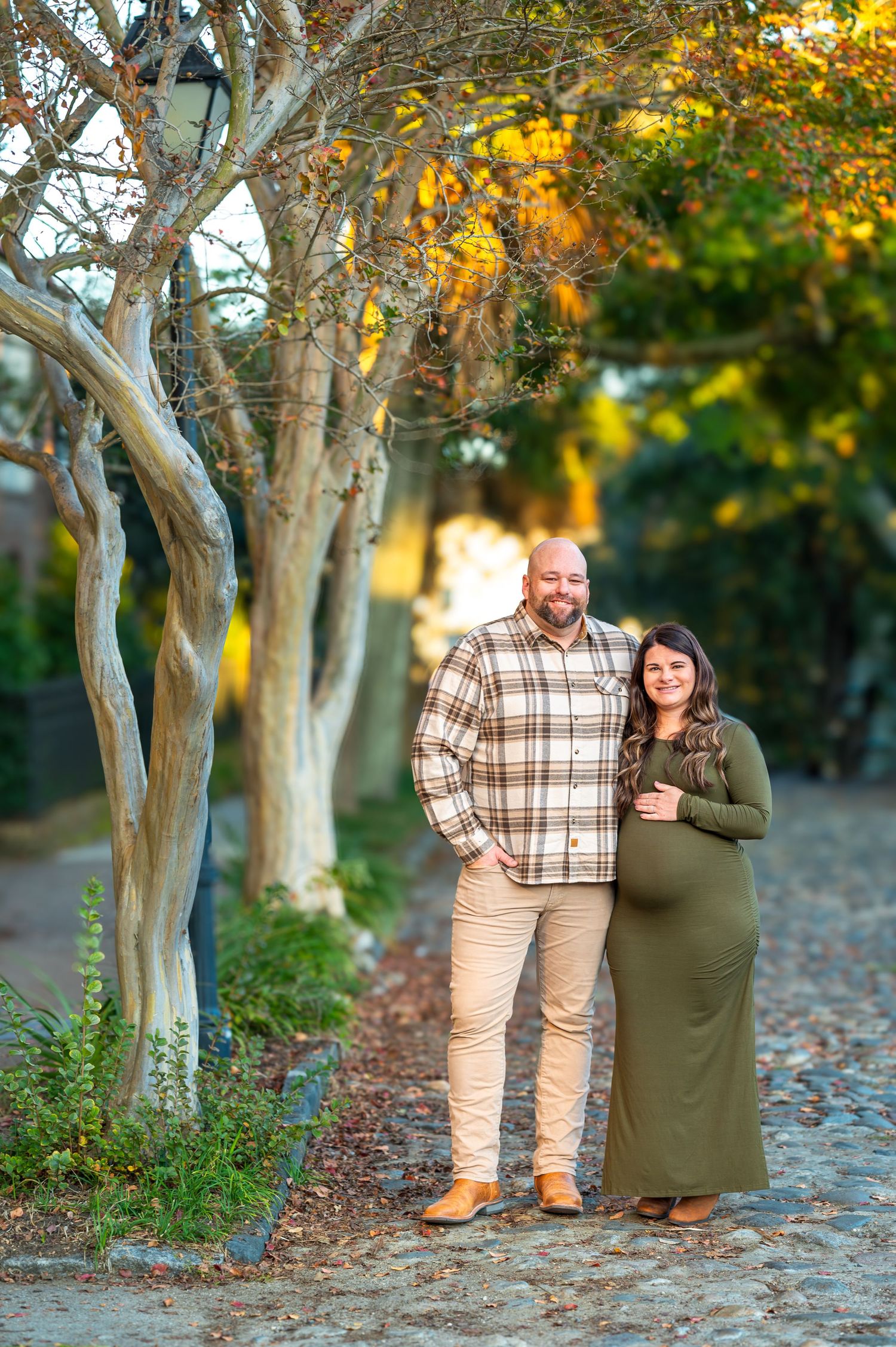 Luna and her husband Aaron sit under a tree with fall foliage on a cobblestone street during their maternity session