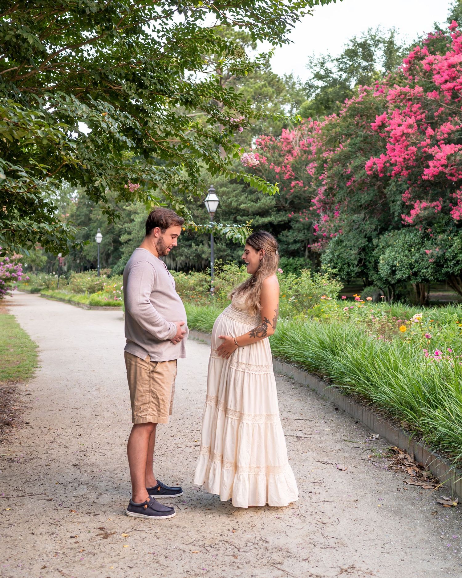 A couple stands facing each other on a garden path with blooming pink flowers.