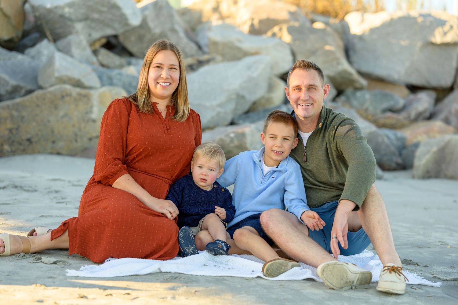 Family with two young boys sitting on beach with rocks behind them