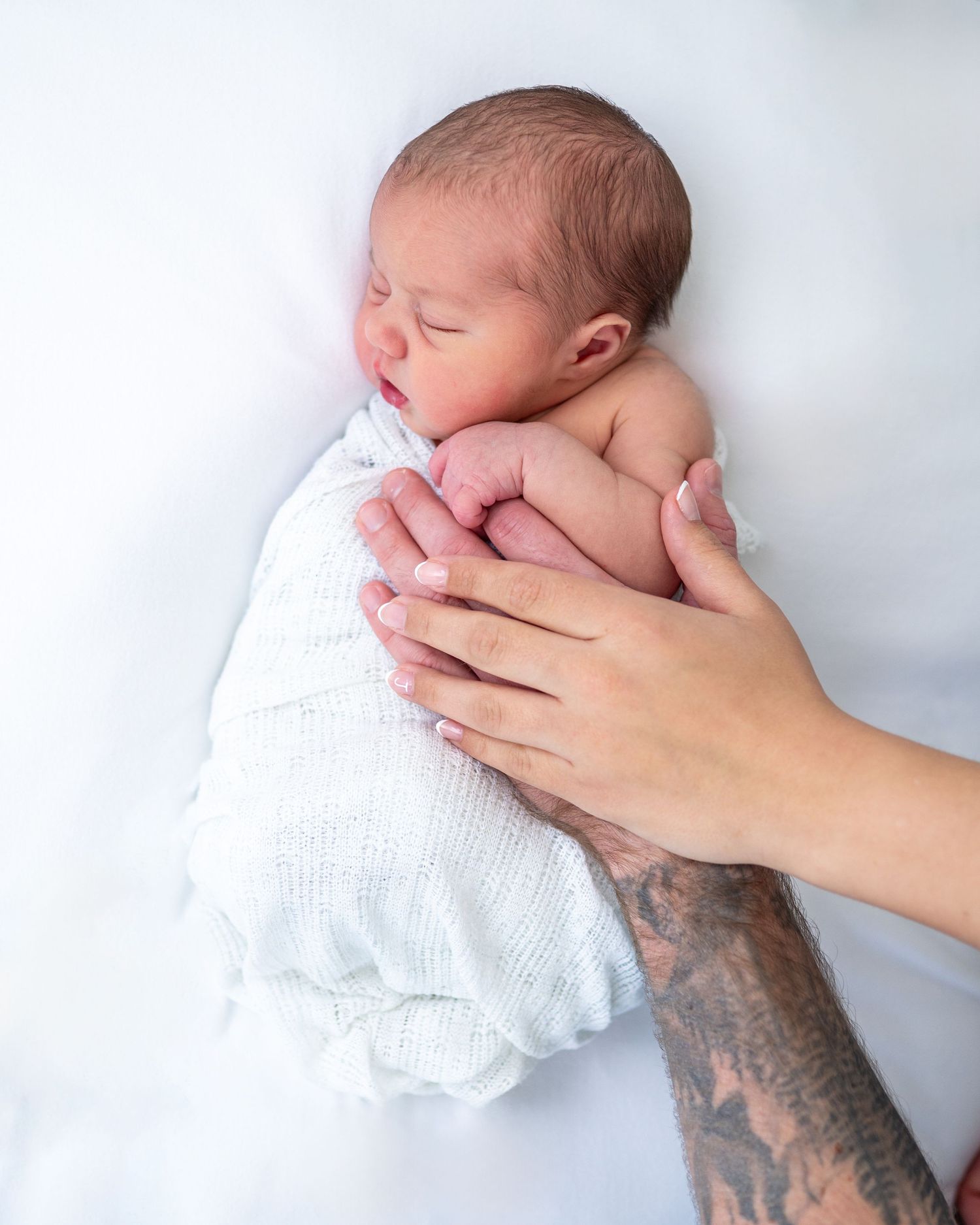 Tattooed hands gently cradling a sleeping newborn baby wrapped in a soft white knit swaddle blanket.
