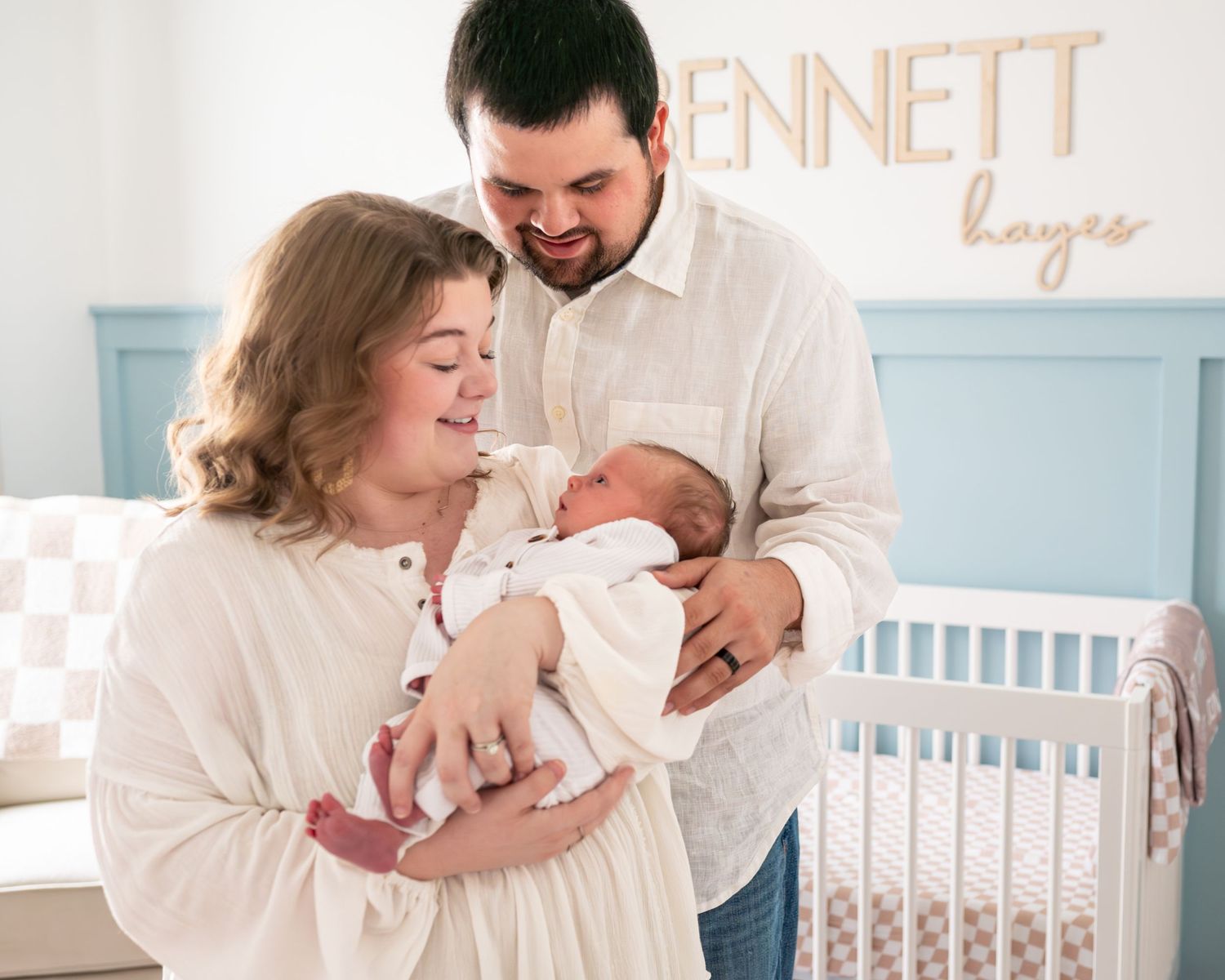 Parents share a tender moment with their newborn baby in a nursery with light blue walls and gold name decor.