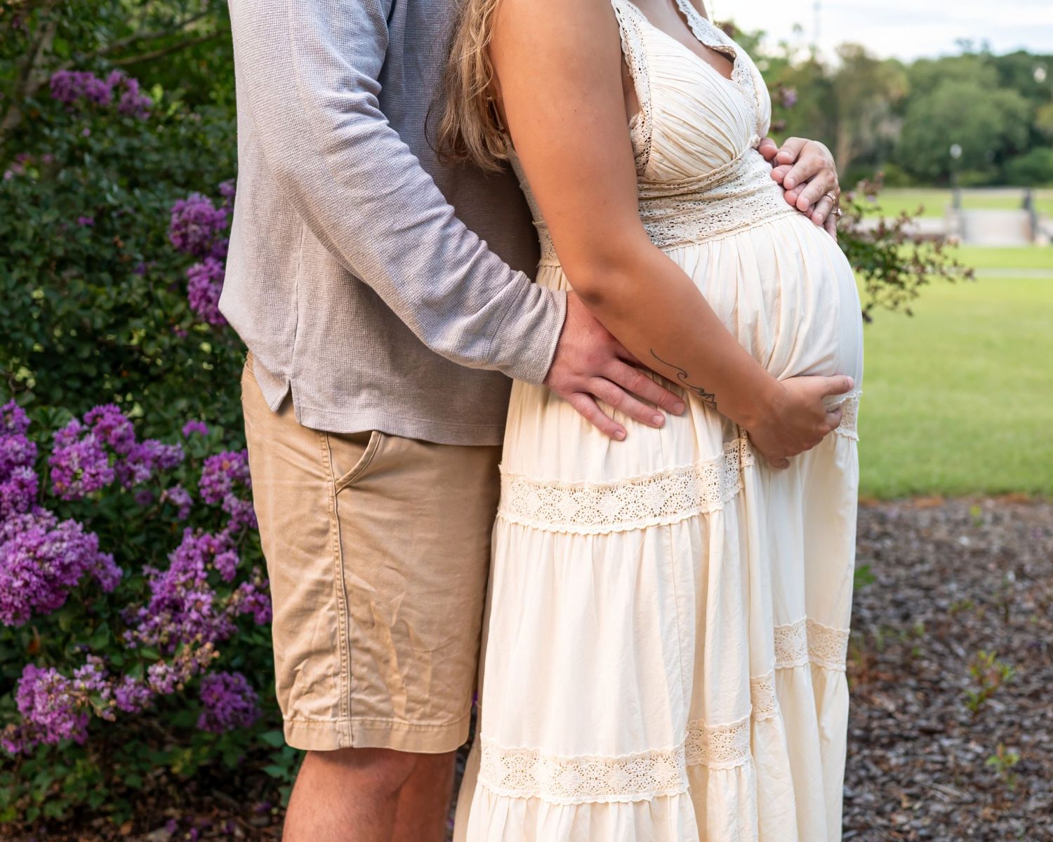 Close-up of an expectant couple embracing in a garden with purple flowers in the background.