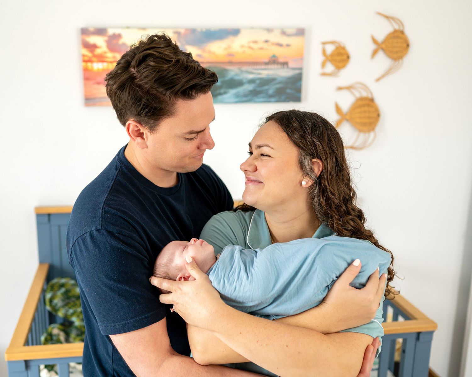 Happy new parents smile at each other while cradling their newborn baby in a coastal-themed nursery.