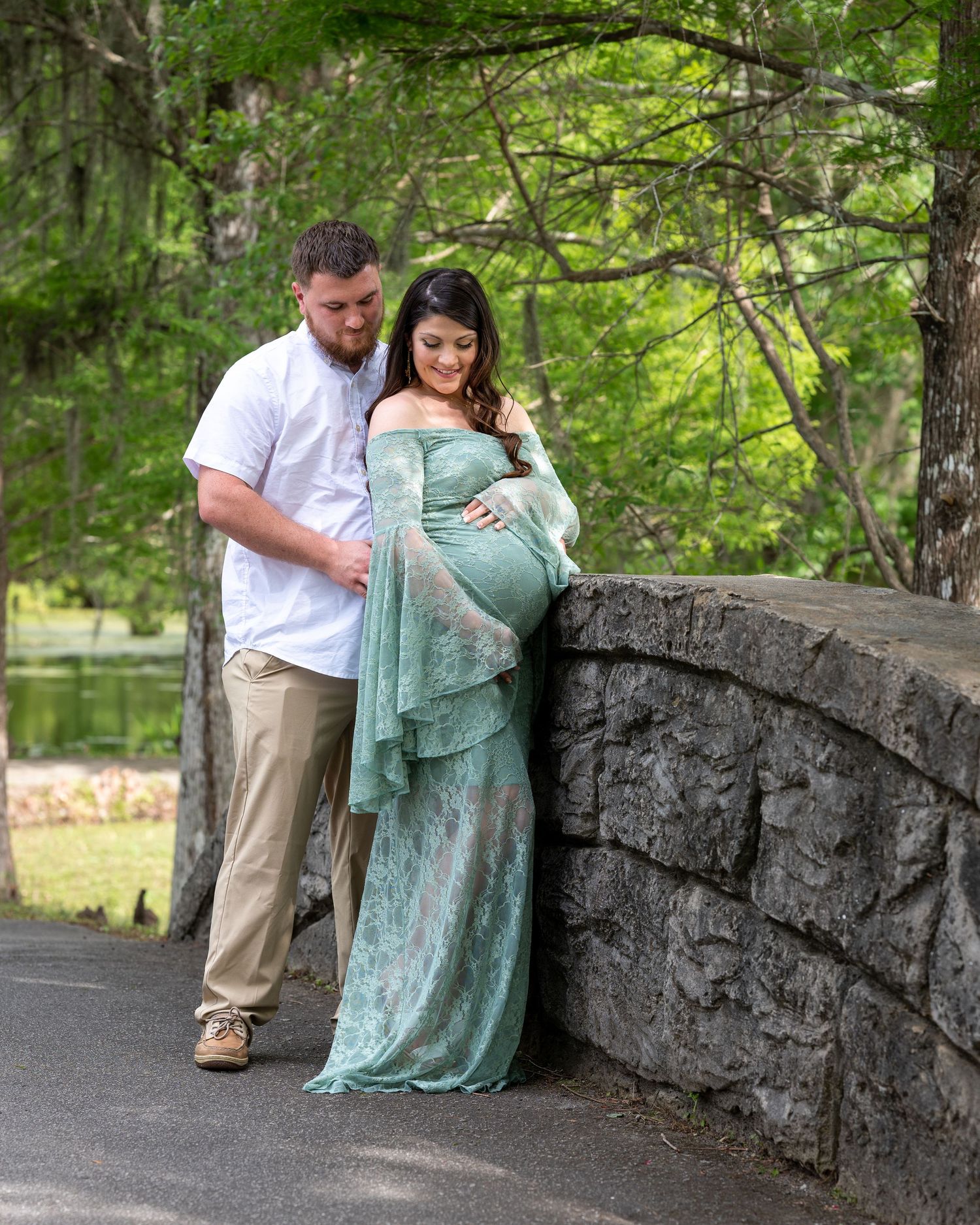 Megan and her husband Brian stand along a bridge, Brian hugs Megan from behind and Megan looks down at her pregnant bump