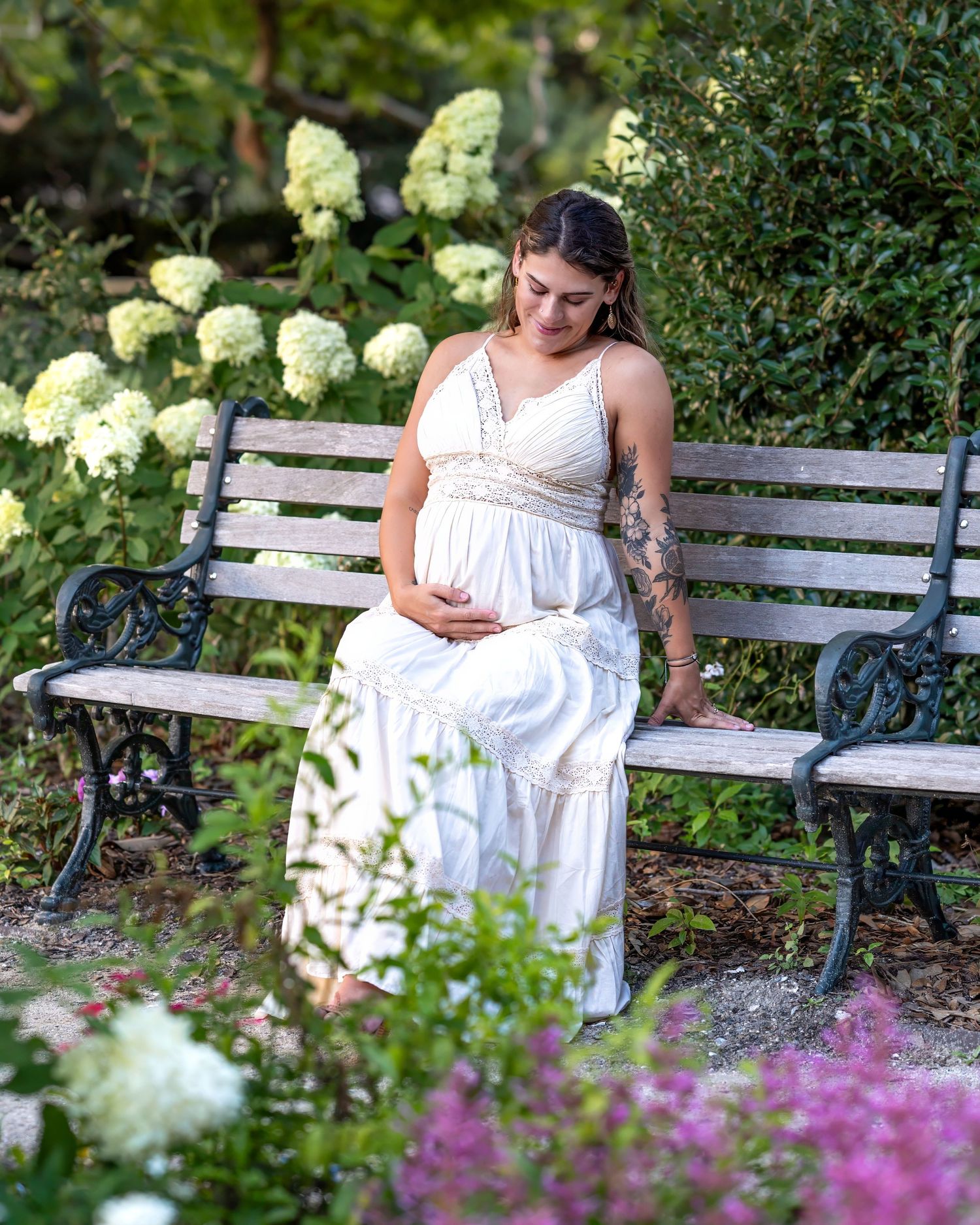 An expectant mother in a white dress sits peacefully on a garden bench surrounded by hydrangeas.