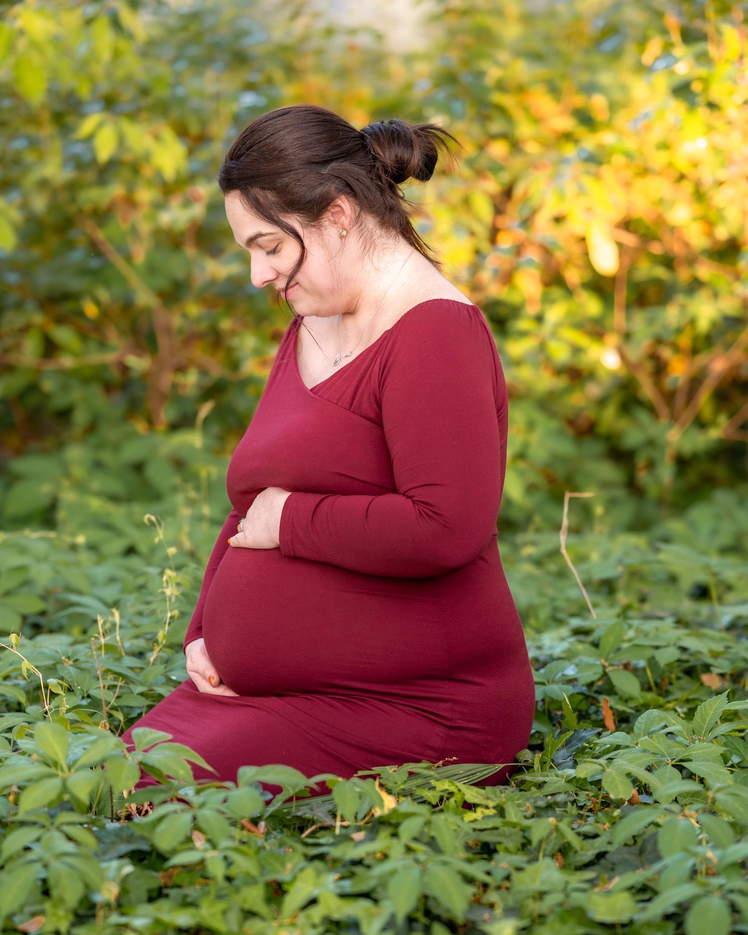 An expectant person in a burgundy dress cradles their belly while sitting in a garden setting.