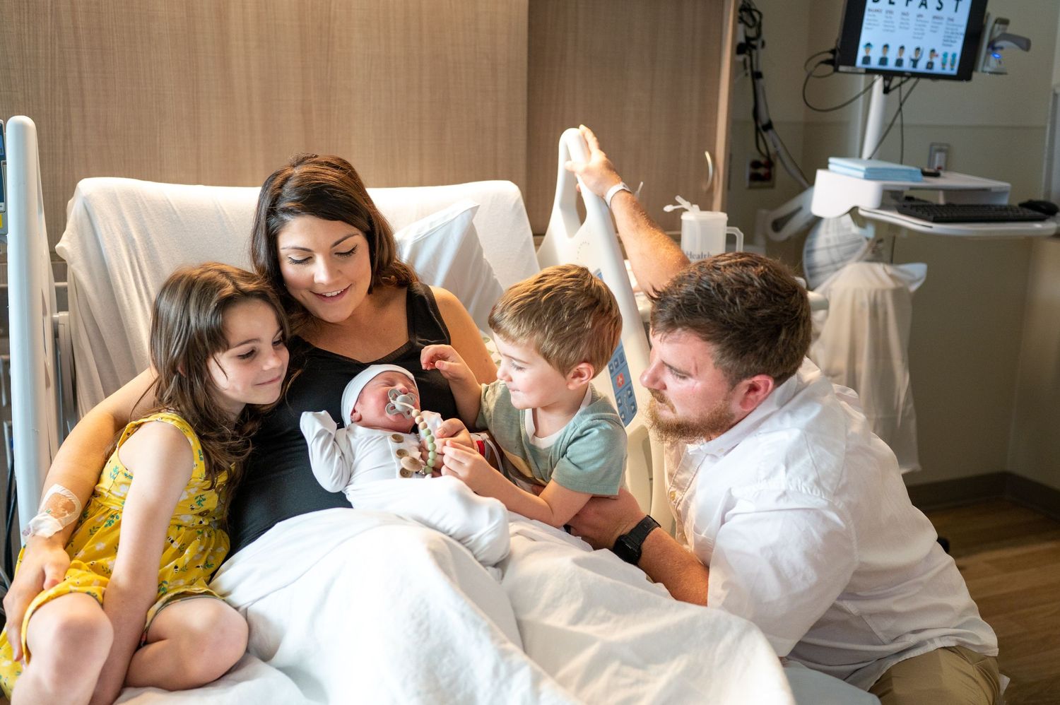 Kayla sits on her hospital bed holding her newborn son with her daughter, son, and husband surrounding them.
