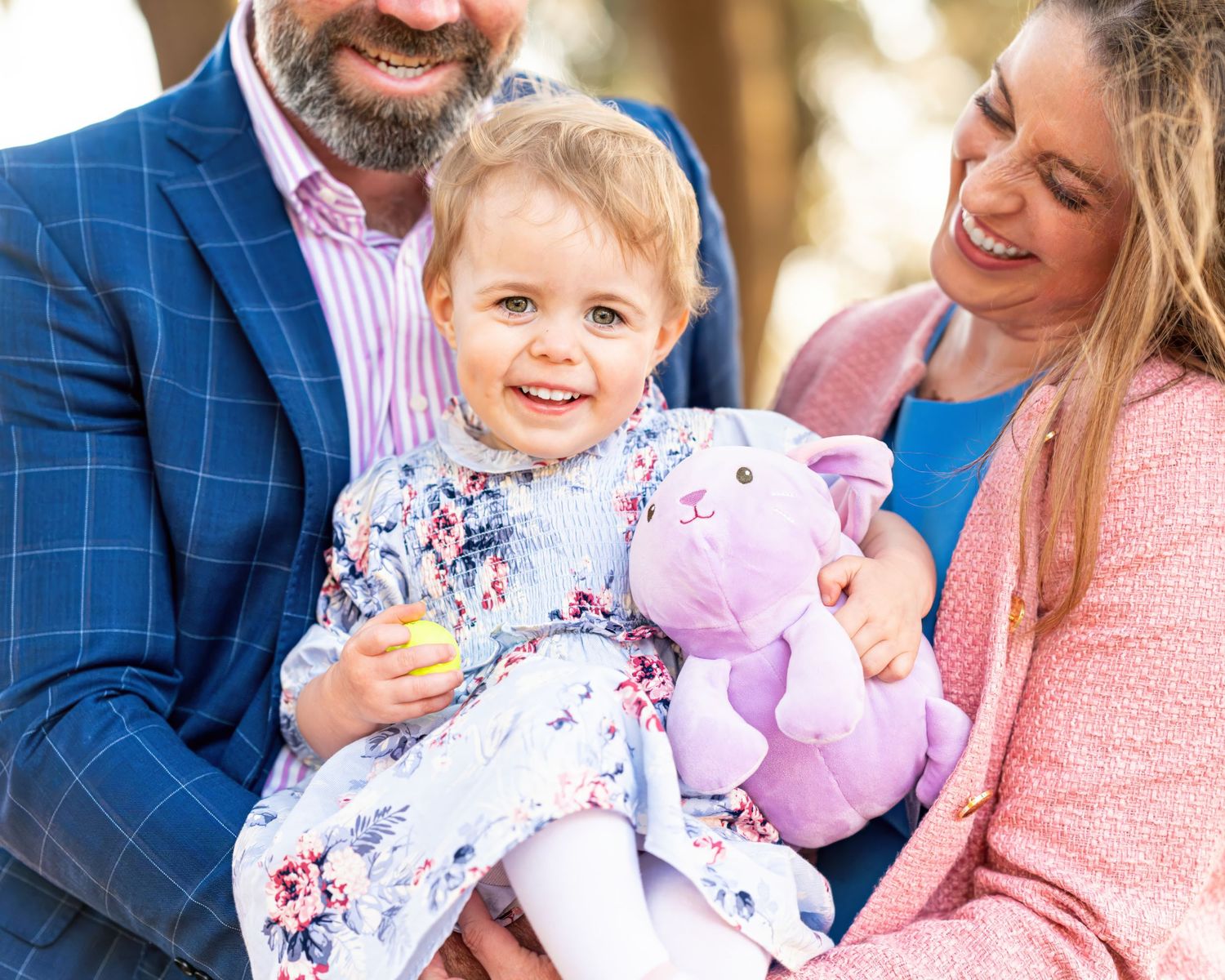 Child in floral dress holds purple teddy bear while outdoors with parents during golden hour photoshoot.
