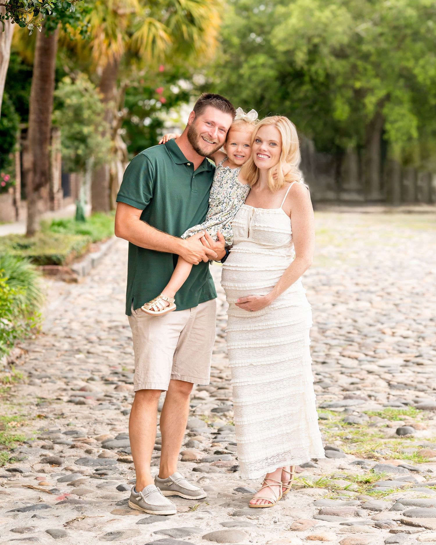 Couple stands together on cobblestone path surrounded by lush greenery on sunny day during their maternity session