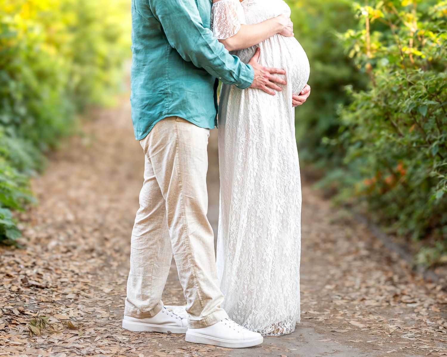 A romantic maternity photo showing a couple embracing in a sunlit garden path.