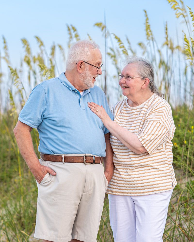 Elderly couple smiling at each other near sea oats on a sunny beach, woman placing hand on man's chest.