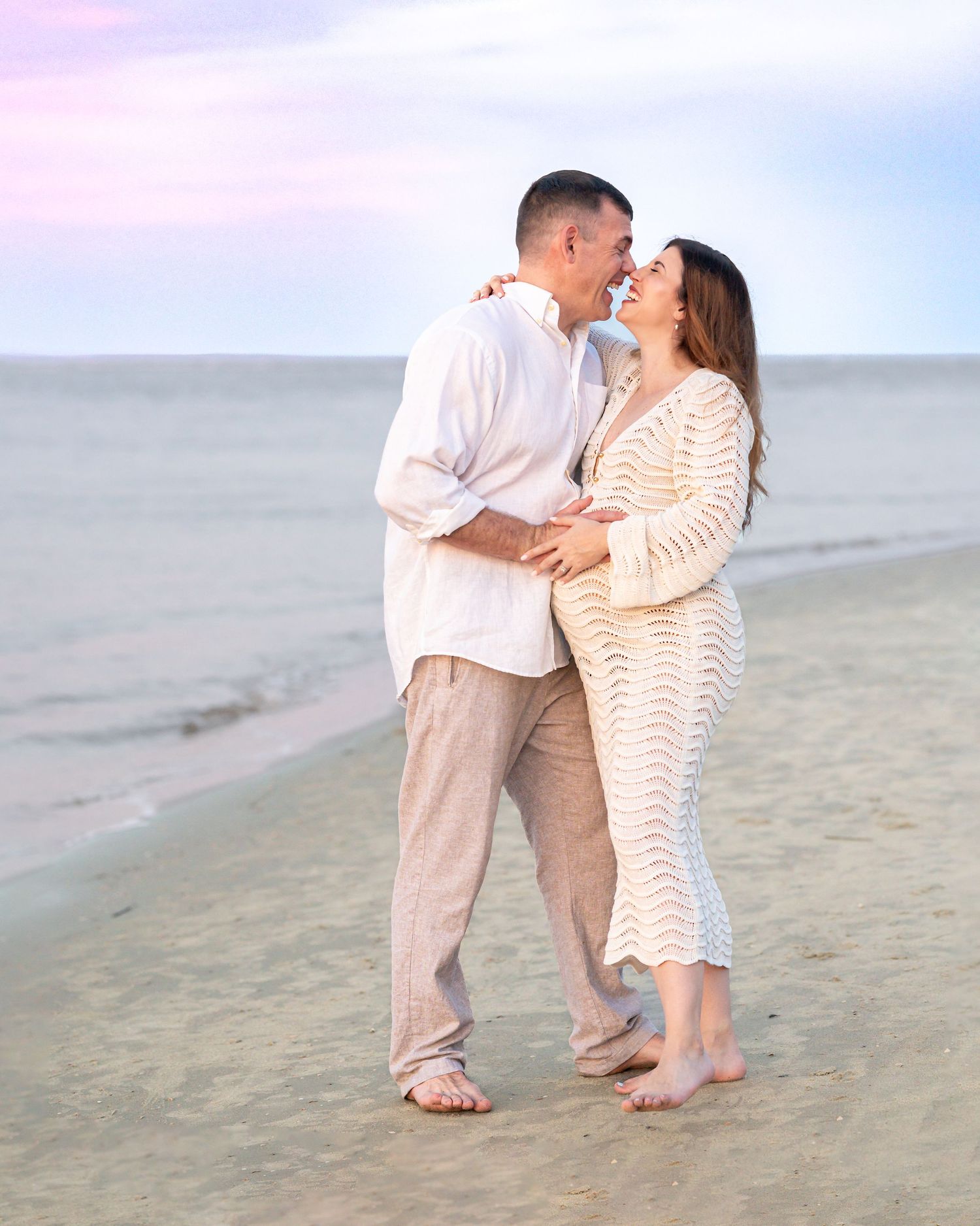 Romantic couple embracing on a sandy beach at sunset, wearing neutral tones, with calm ocean waves in the background.