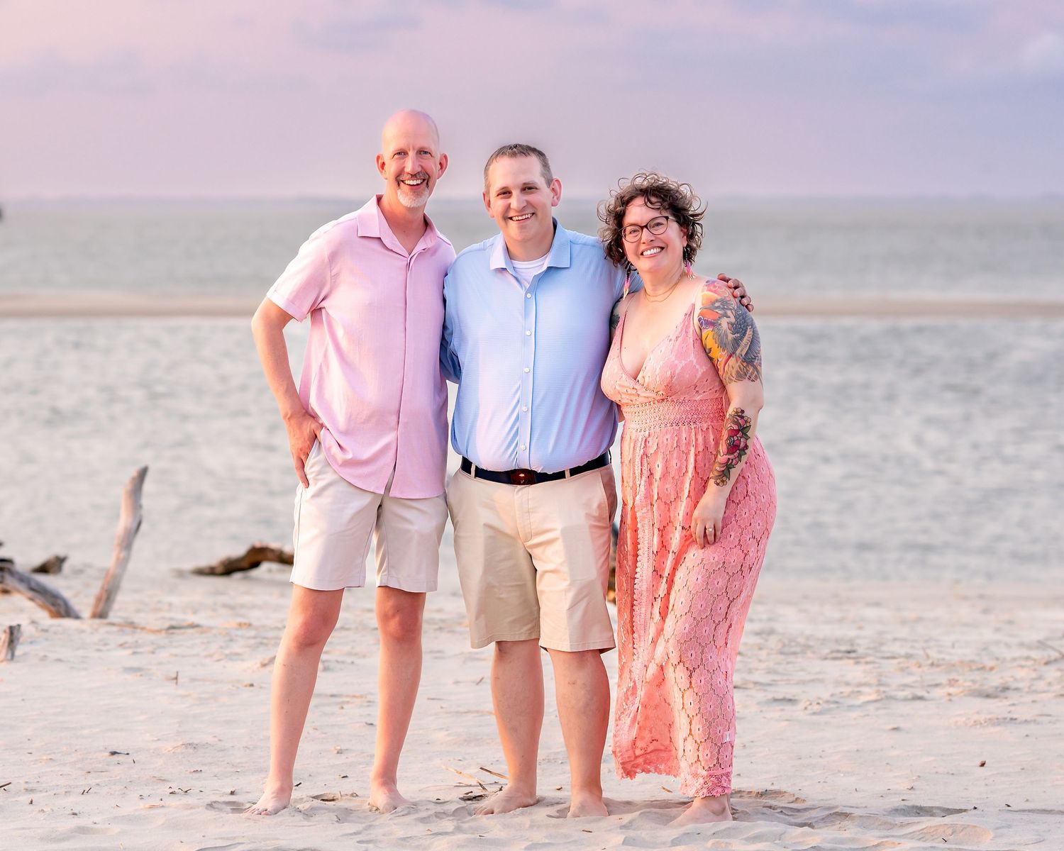 Three people smiling together on a sandy beach at sunset, wearing pastel outfits with ocean waves in the background.