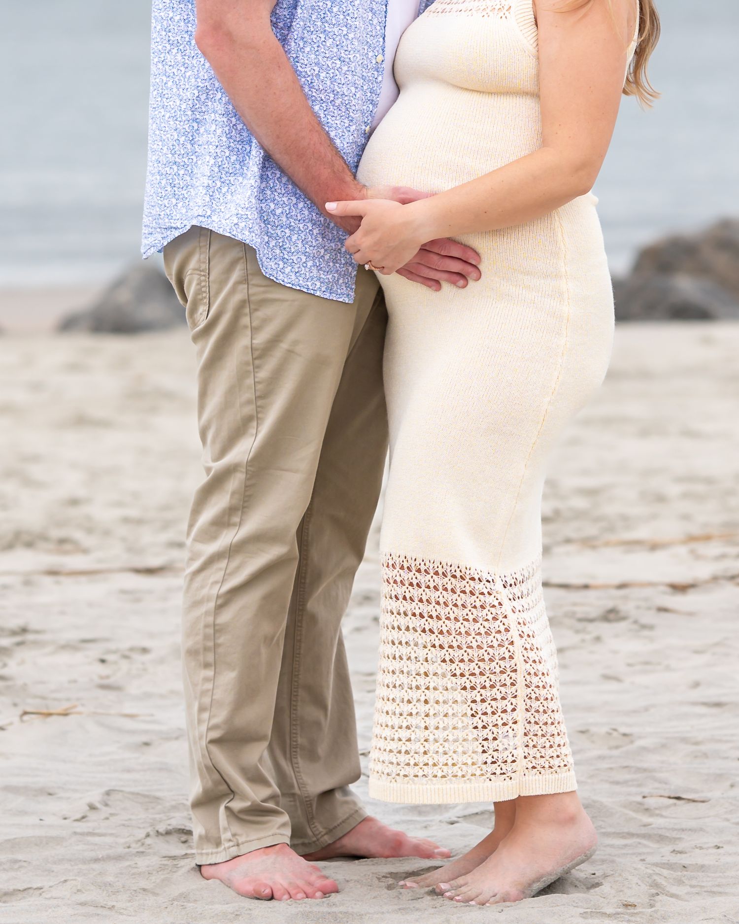 Pregnant couple standing barefoot on a sandy beach, cradling baby bump, wearing casual summer outfits.