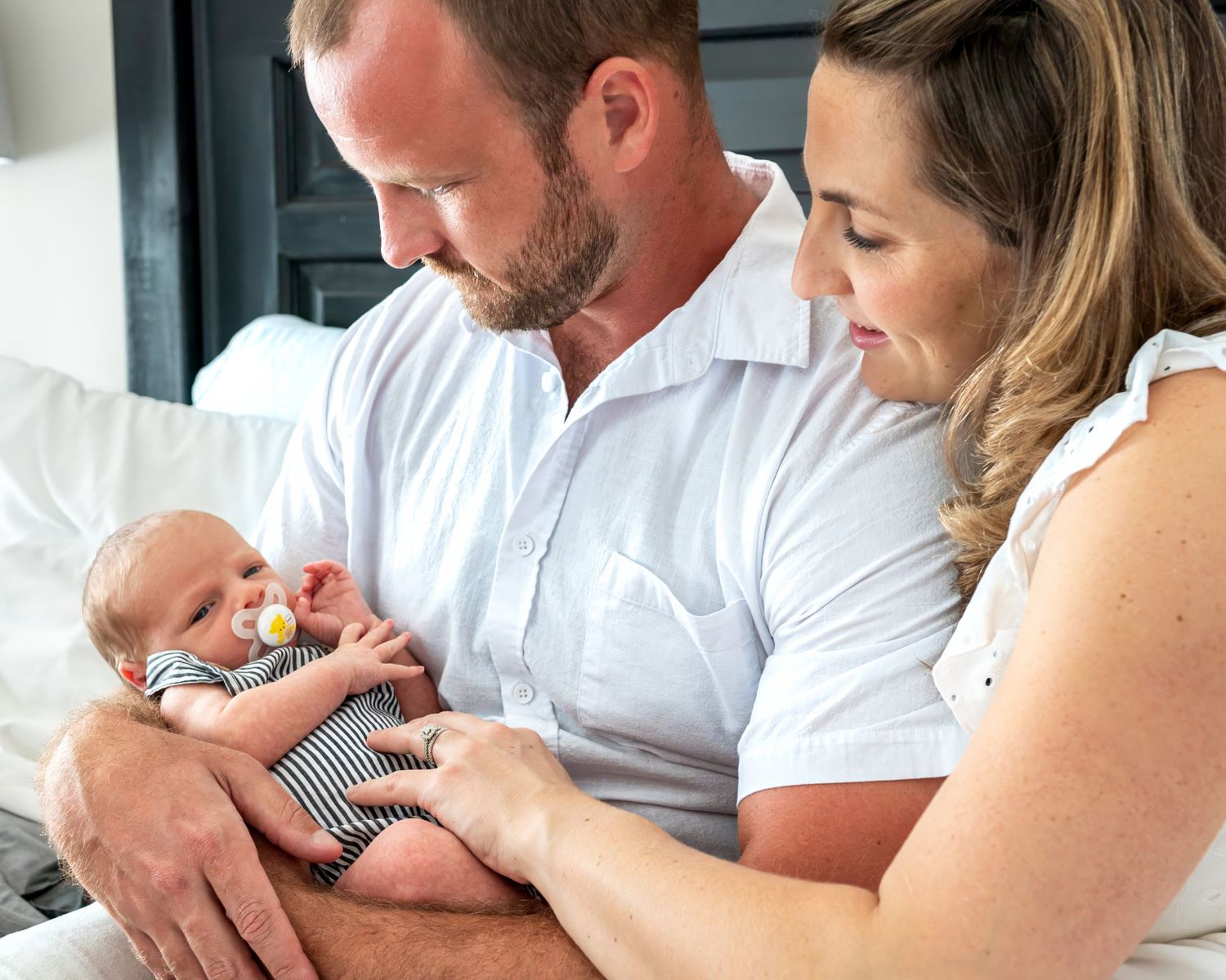 Parents in white clothing bonding with newborn baby wearing striped onesie.