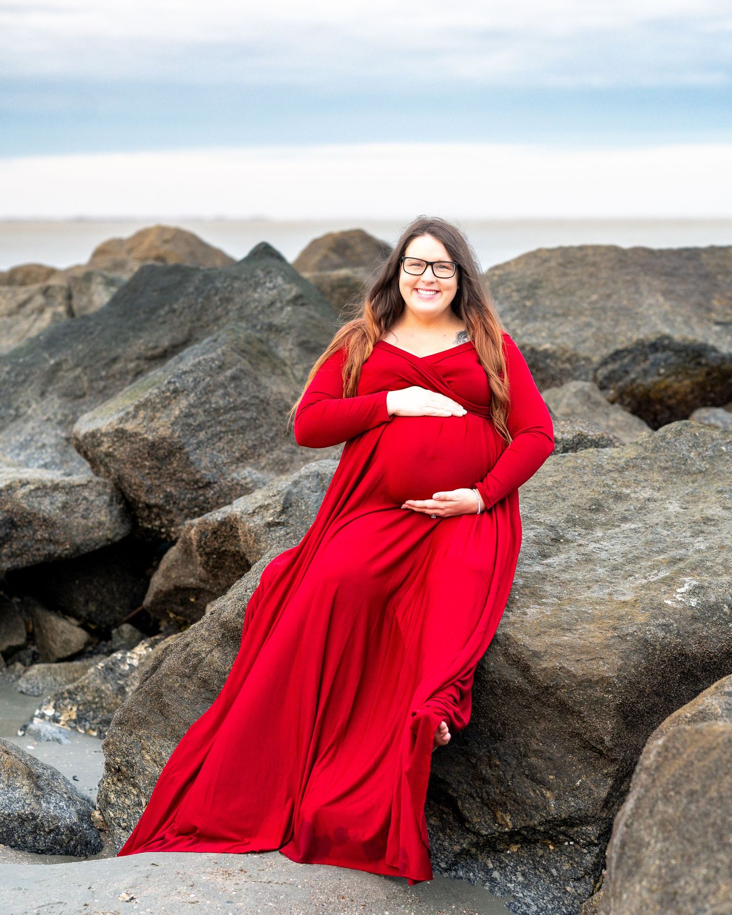 A person in a flowing red maternity dress poses on coastal rocks during a beach photoshoot at sunset.