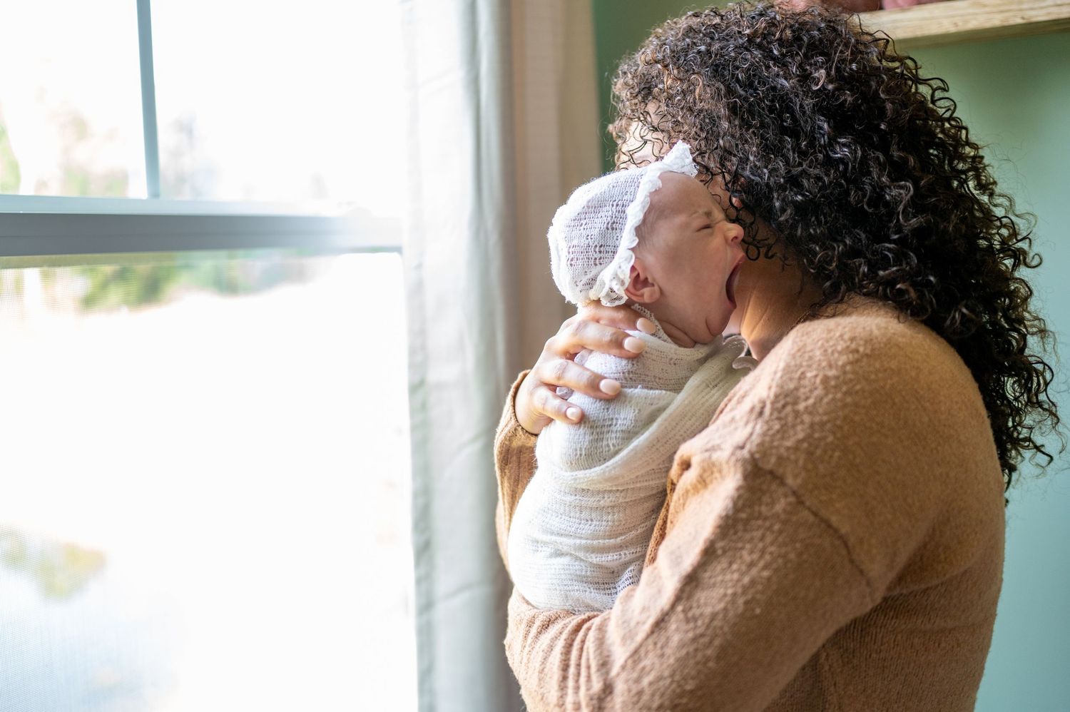 A newborn little girl in a bonnet is being held on her mother's shoulders as she yawns a big yawn