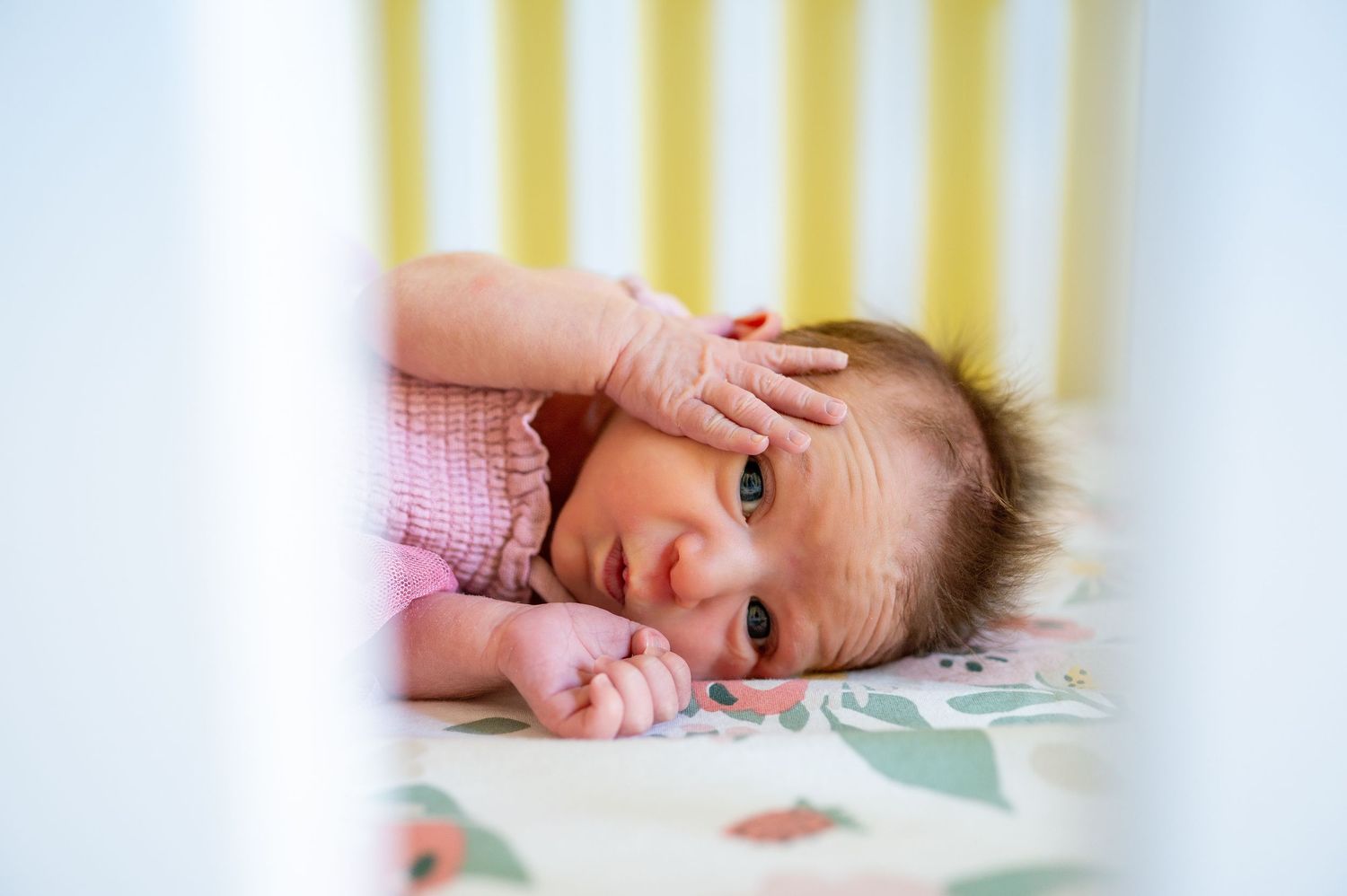 Newborn baby resting in white crib with yellow striped wall behind.