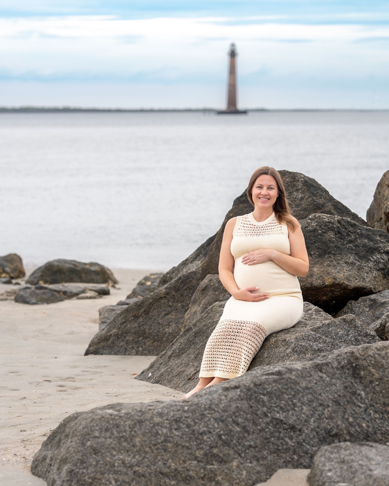 Pregnant woman in cream crochet dress sitting on rocks at beach with lighthouse in background.