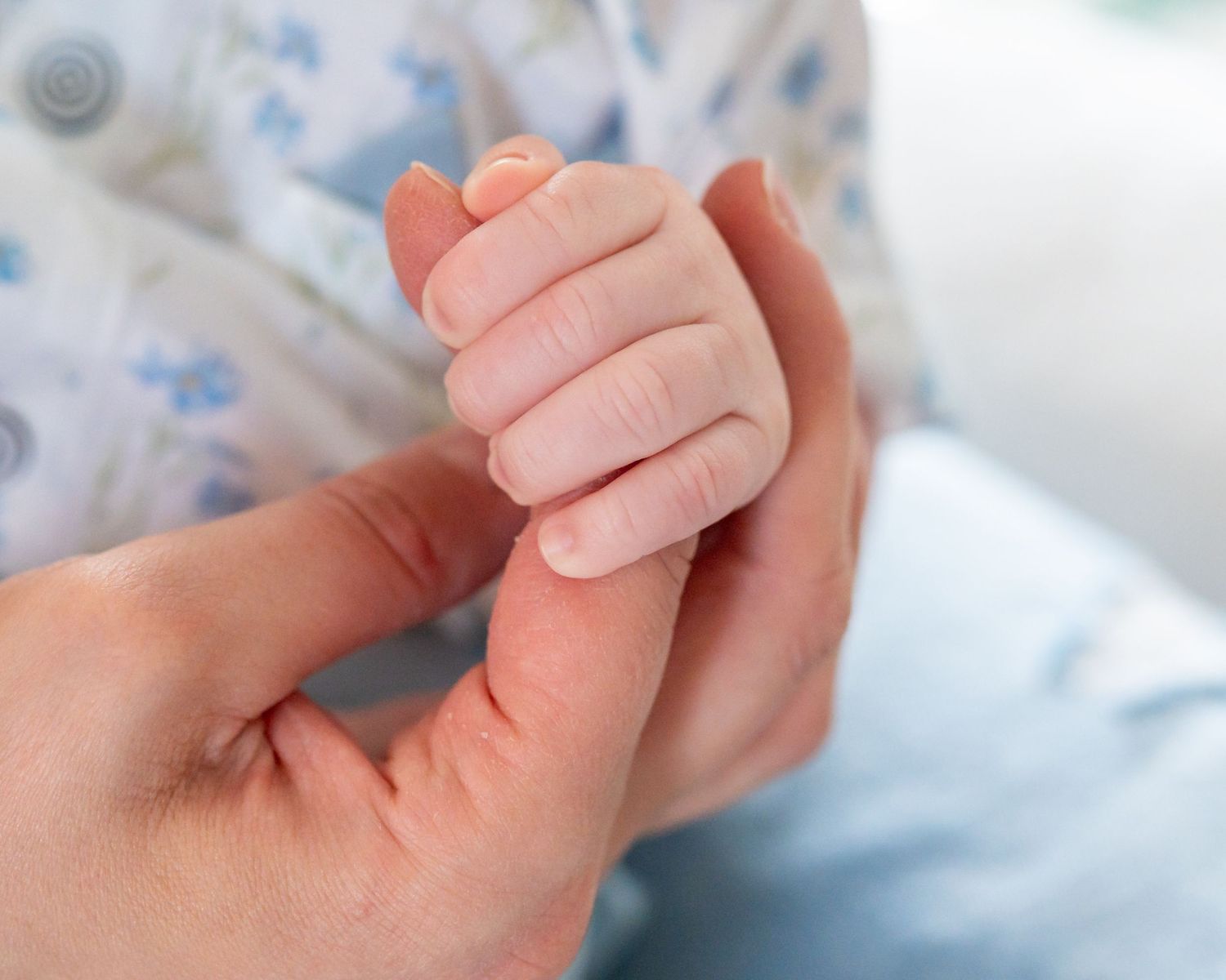 Close-up view of a tiny newborn baby hand gently holding an adult's finger against soft blue blanket background.