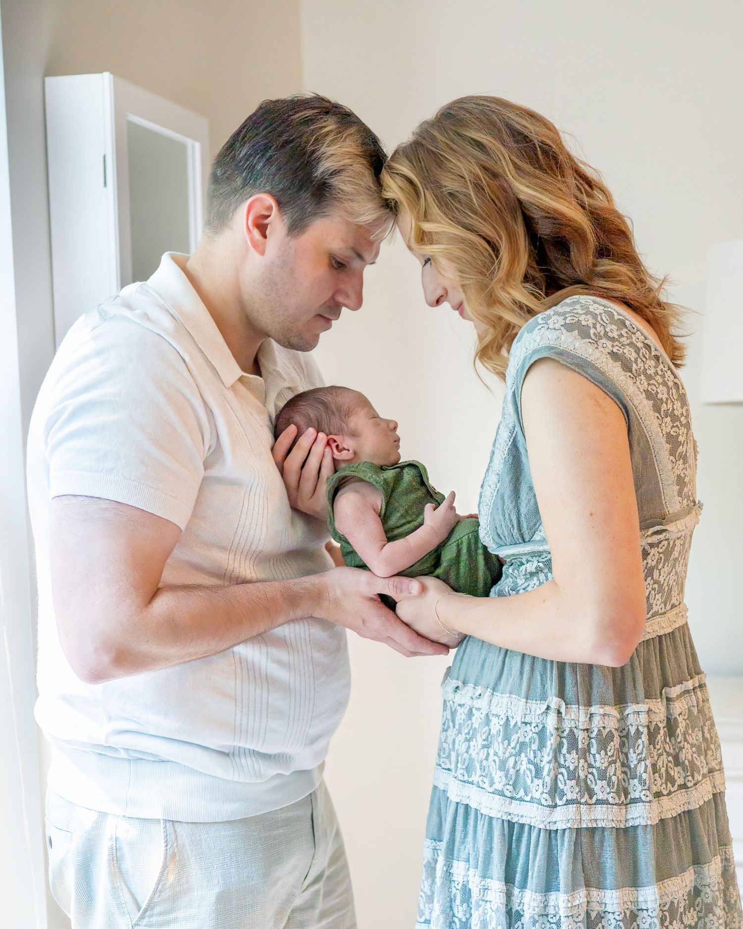 New parents tenderly hold their newborn baby together, foreheads touching, in a soft natural light indoor setting.