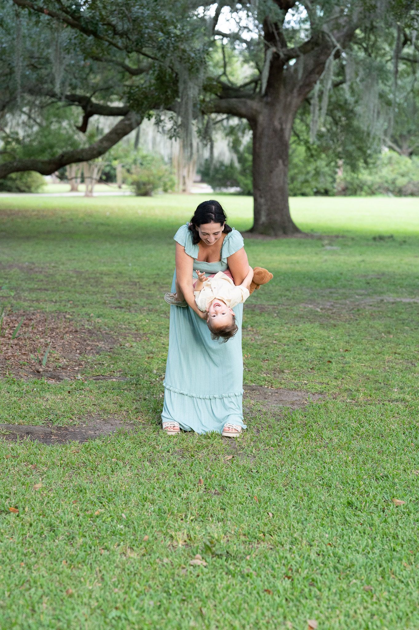 Unedited maternity session. Mom in blue dress holds her toddler son upside down as they play in a park.