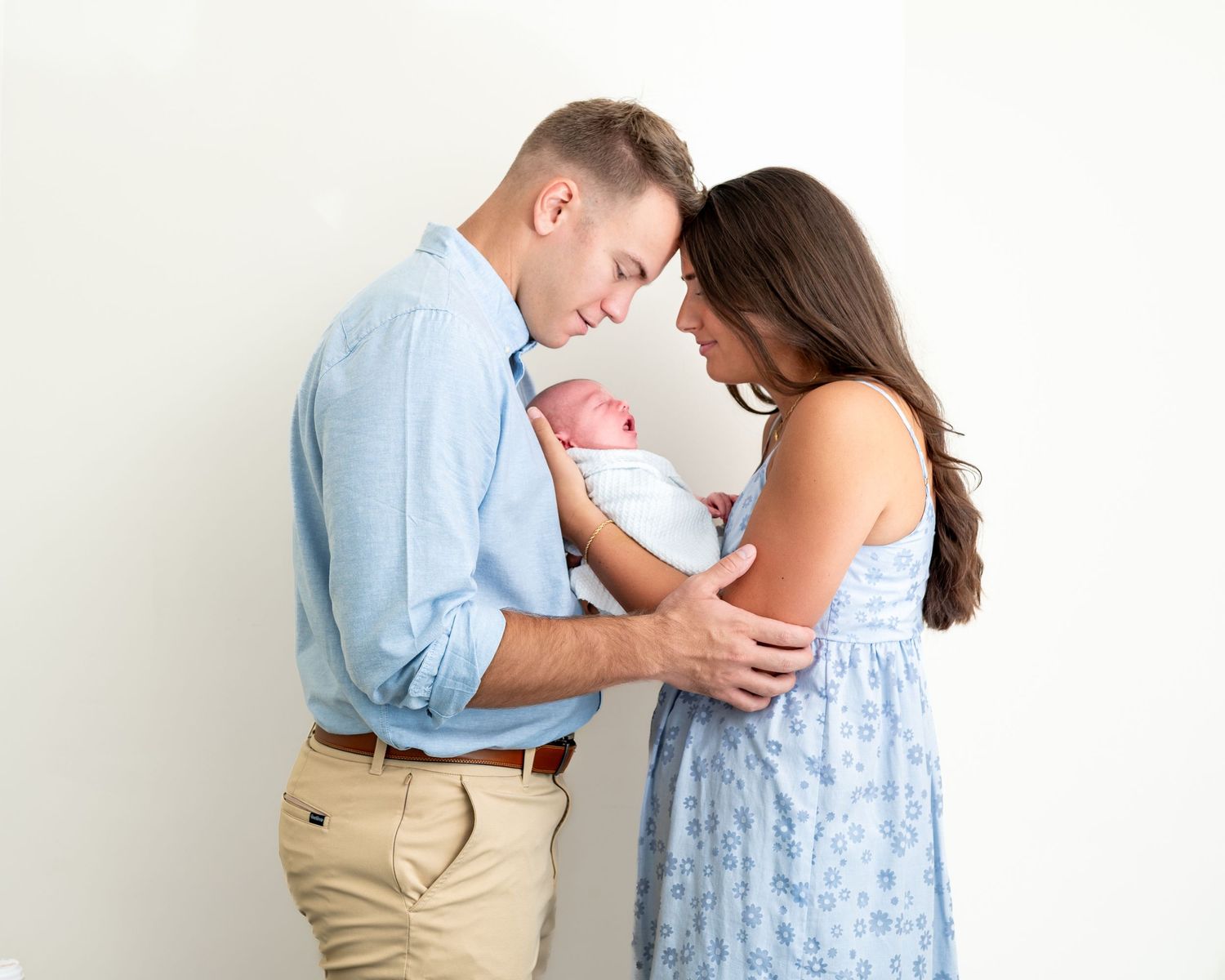 Claire and her husband stand facing each other with their foreheads touching as they hold their newborn son between them.