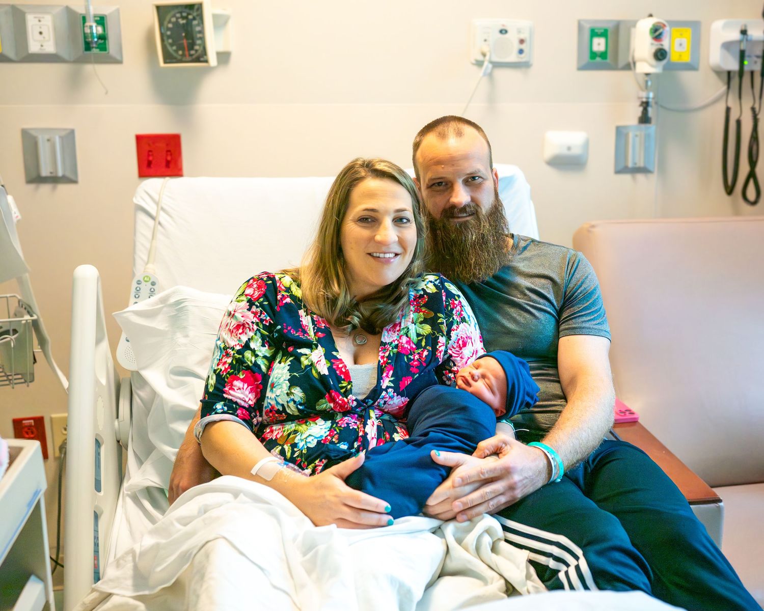New family rests together on hospital bed with baby in blue swaddle.