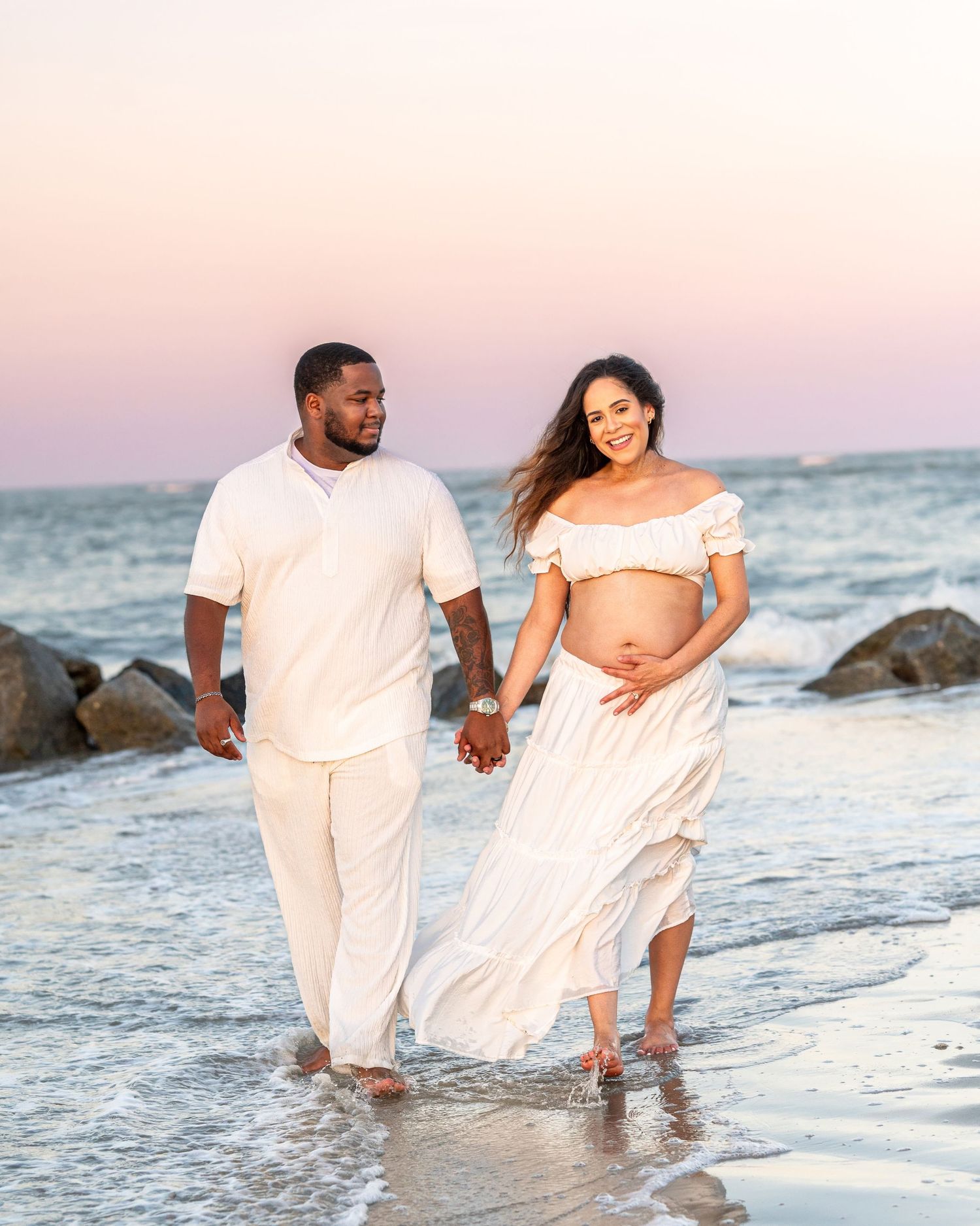 Romantic beach photoshoot with couple walking in white outfits along the shoreline.