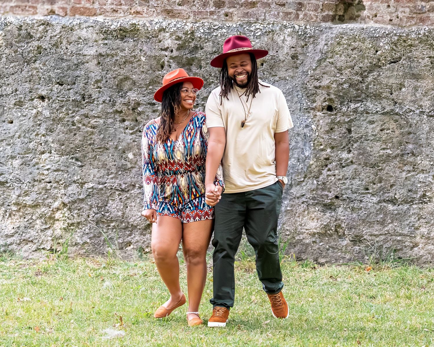 Romantic couple in casual attire relaxing against historic stone wall backdrop.