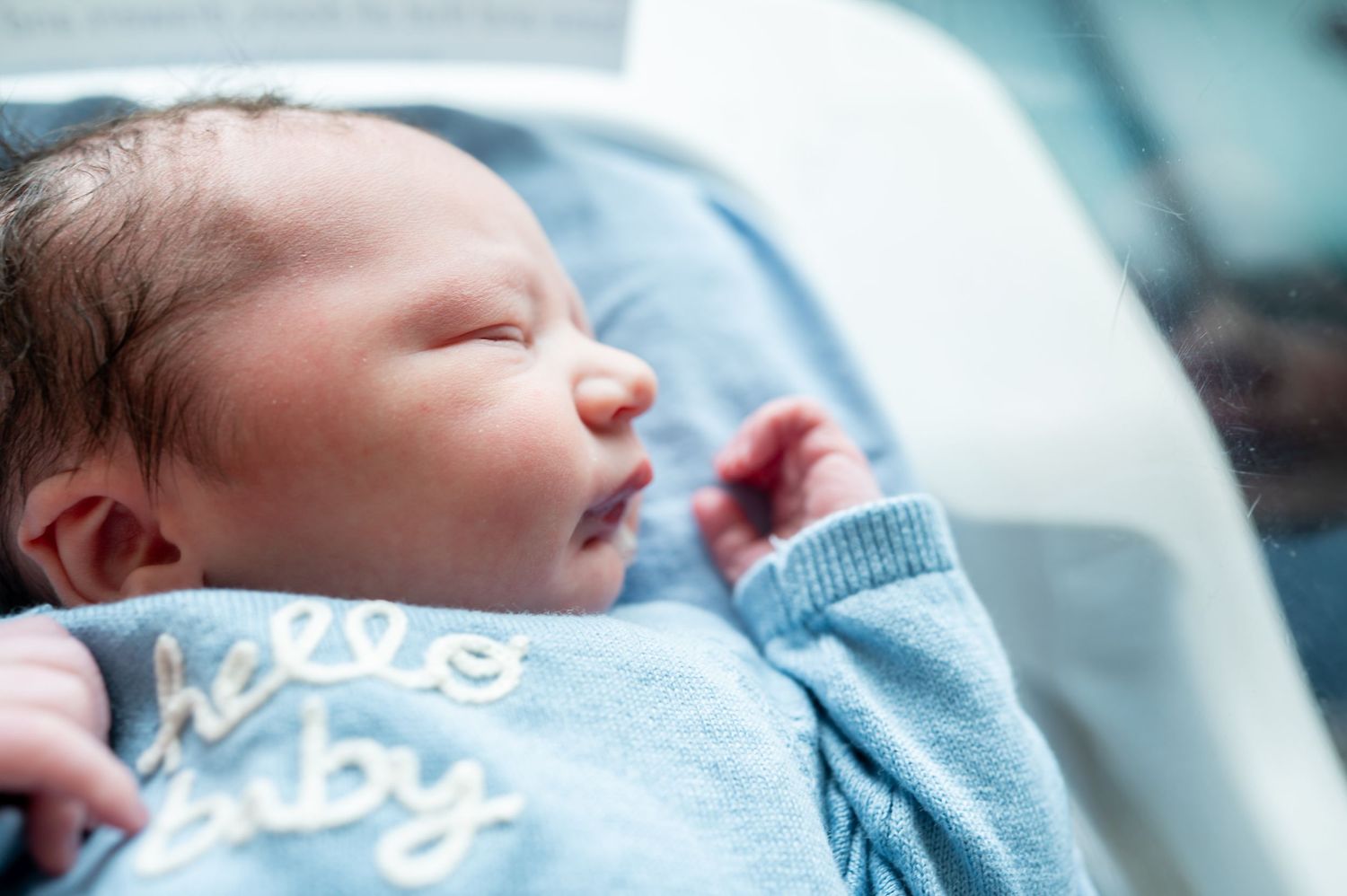 Newborn baby sleeping peacefully in a blue knit sweater that reads hello baby, wrapped in a soft blanket.