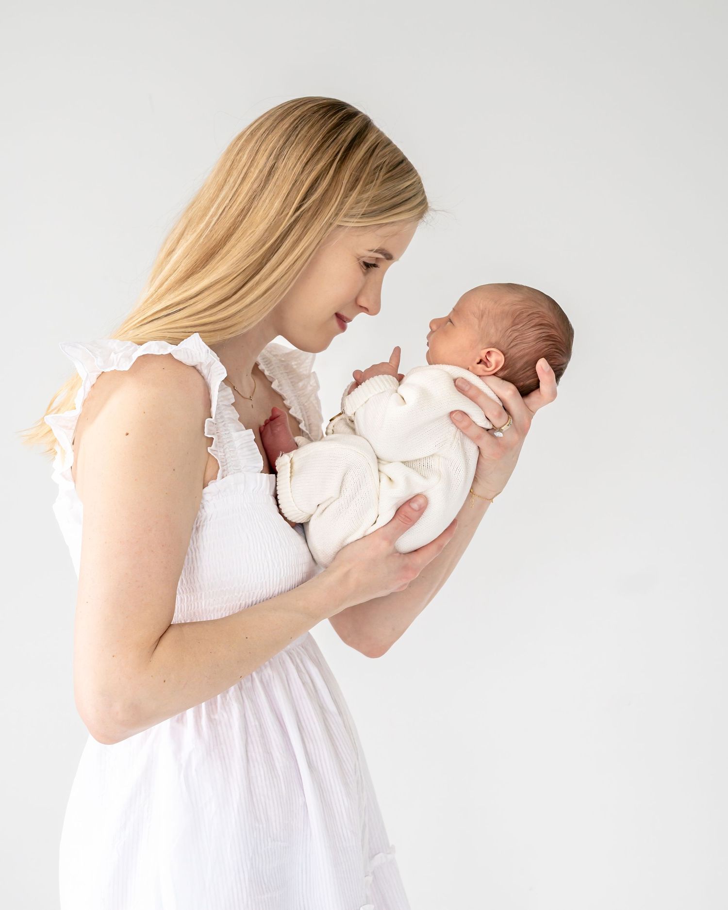 A tender moment between parent and newborn baby both wearing white against a light gray background.
