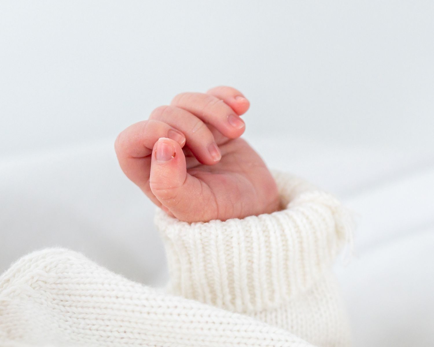 A tiny baby hand reaches out from a white knitted sweater sleeve against a light background.