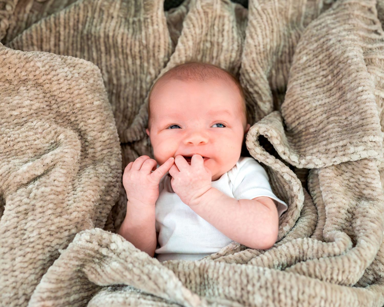 A happy baby wrapped in a cozy gray blanket smiles while chewing on their fingers.