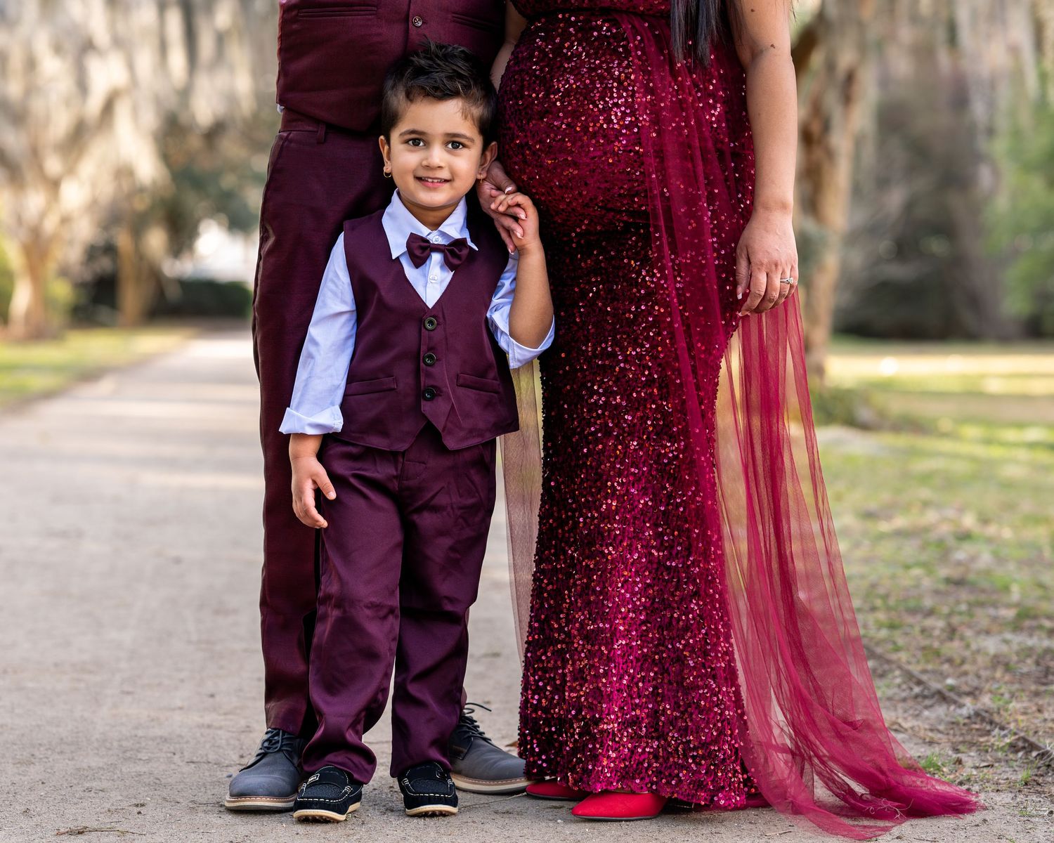 Sandra and her husband stand side by side holding their toddler son's hand.  Close up of the toddler without seeing the faces of parents