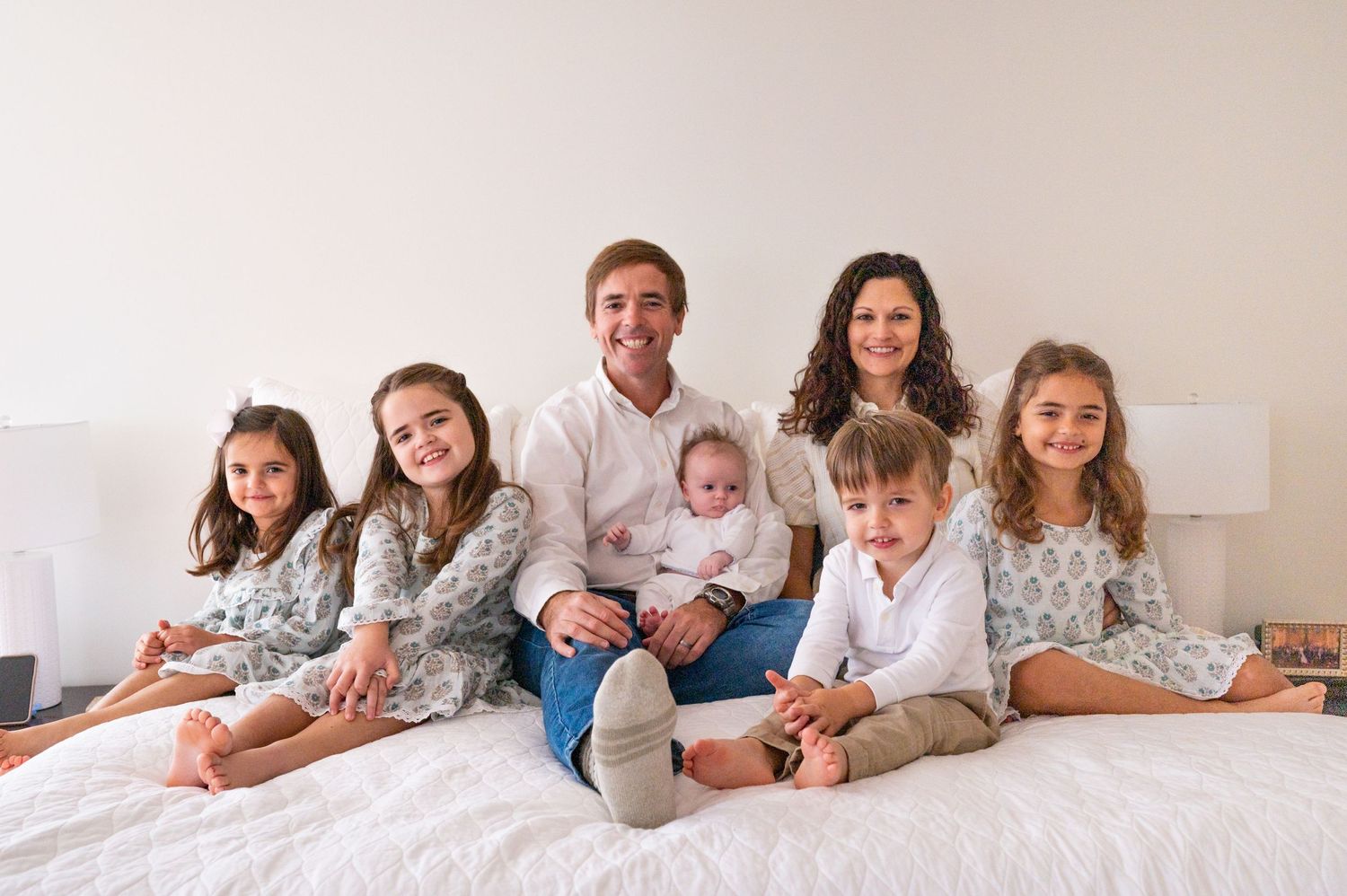 A large family with four young children and a newborn pose on the parent's bed.