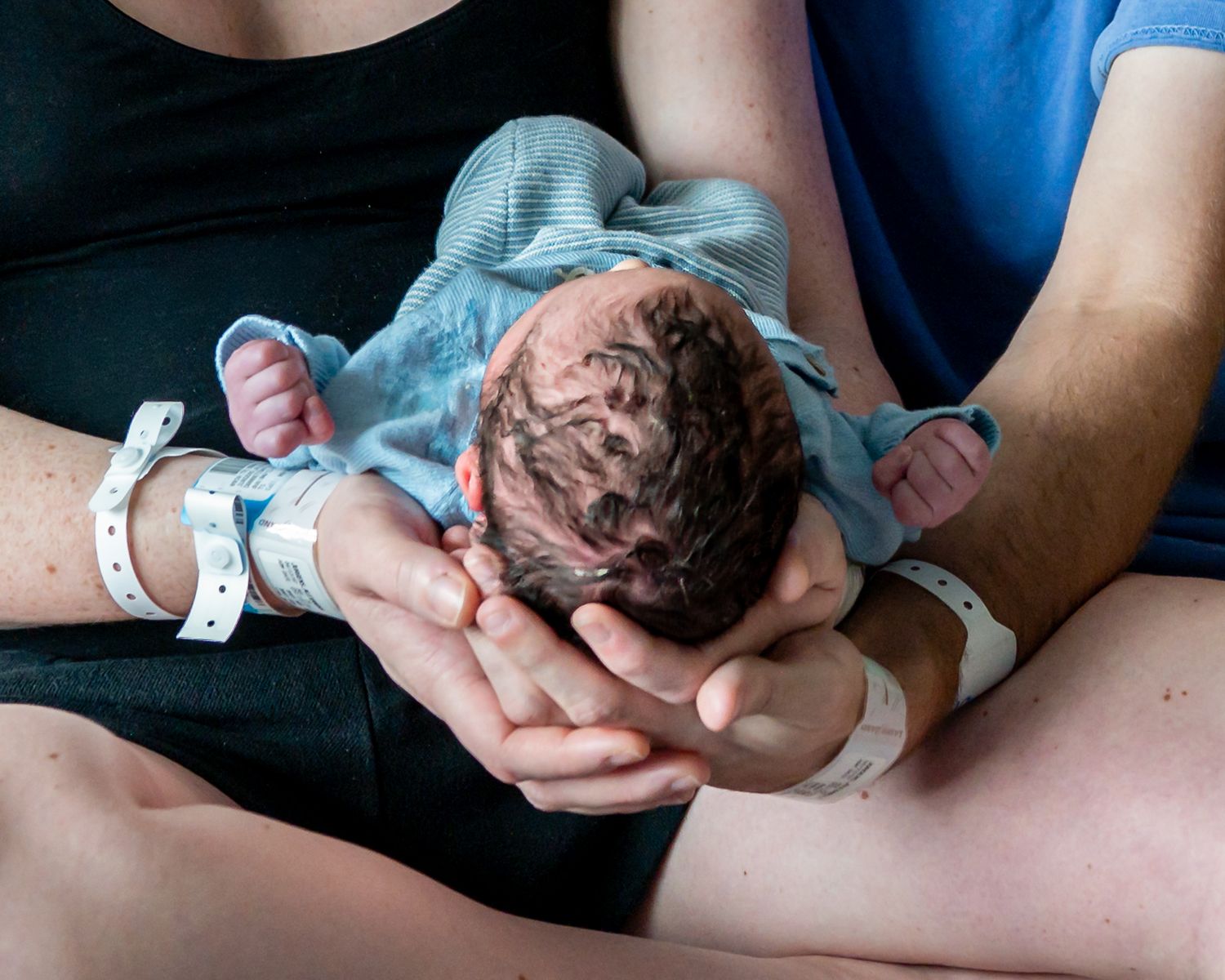 New parents cradling their newborn baby in a hospital room, both wearing identification wristbands after birth.