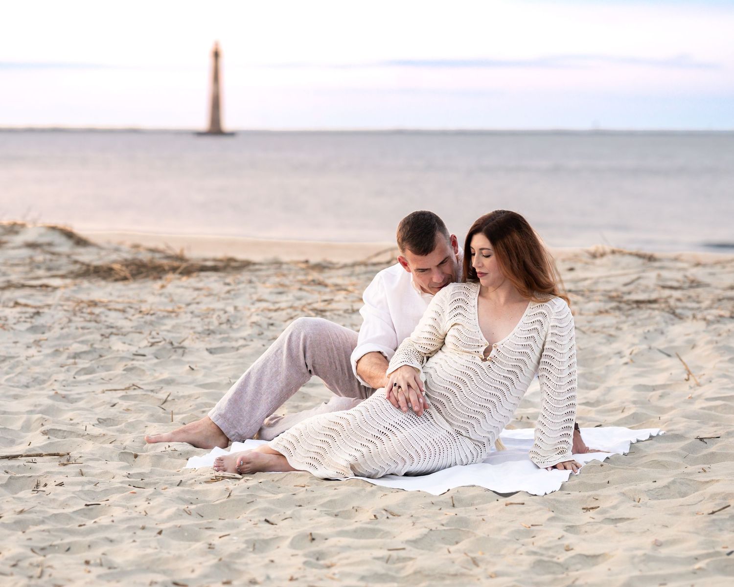Romantic couple sitting on sandy beach blanket on Folly Beach at sunset during their maternity photography session.