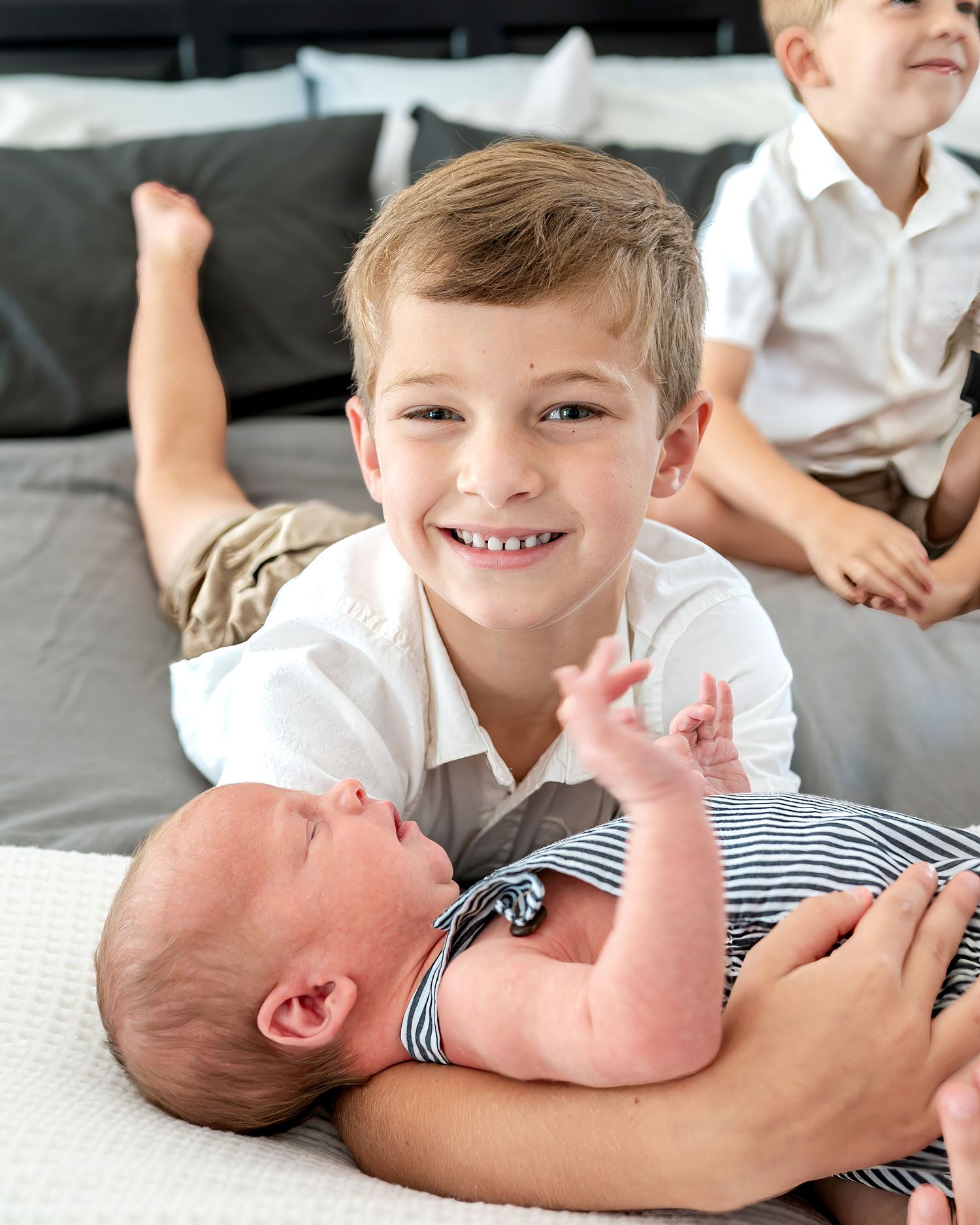 Loving brother holds newborn sibling wearing striped outfit on gray bedding.