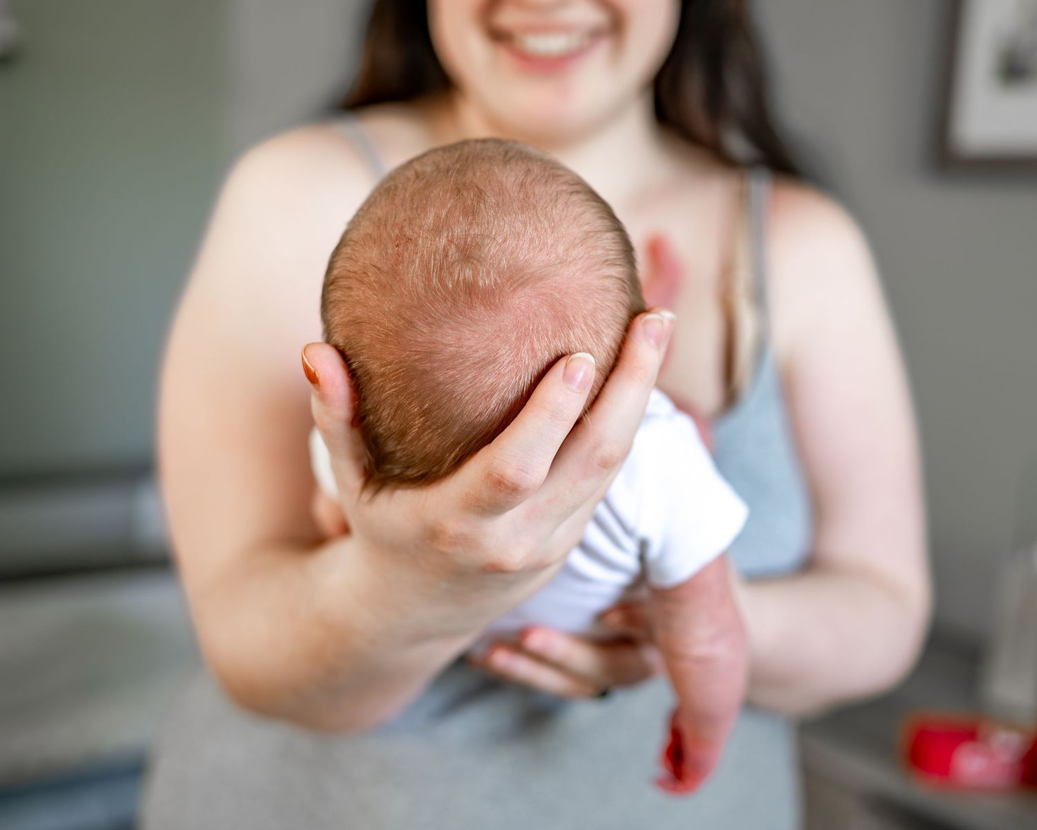 Parent gently cradles newborn baby's head while sitting on gray sofa in living room.
