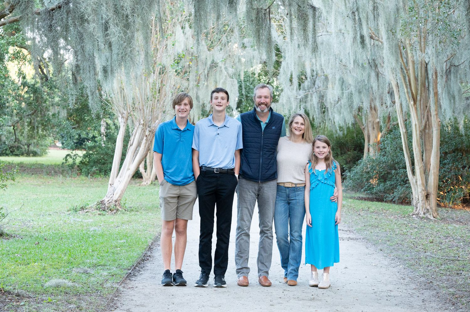 Unedited Photo: A family in coordinated blue and white outfits poses together on a tree-lined path for outdoor portraits.