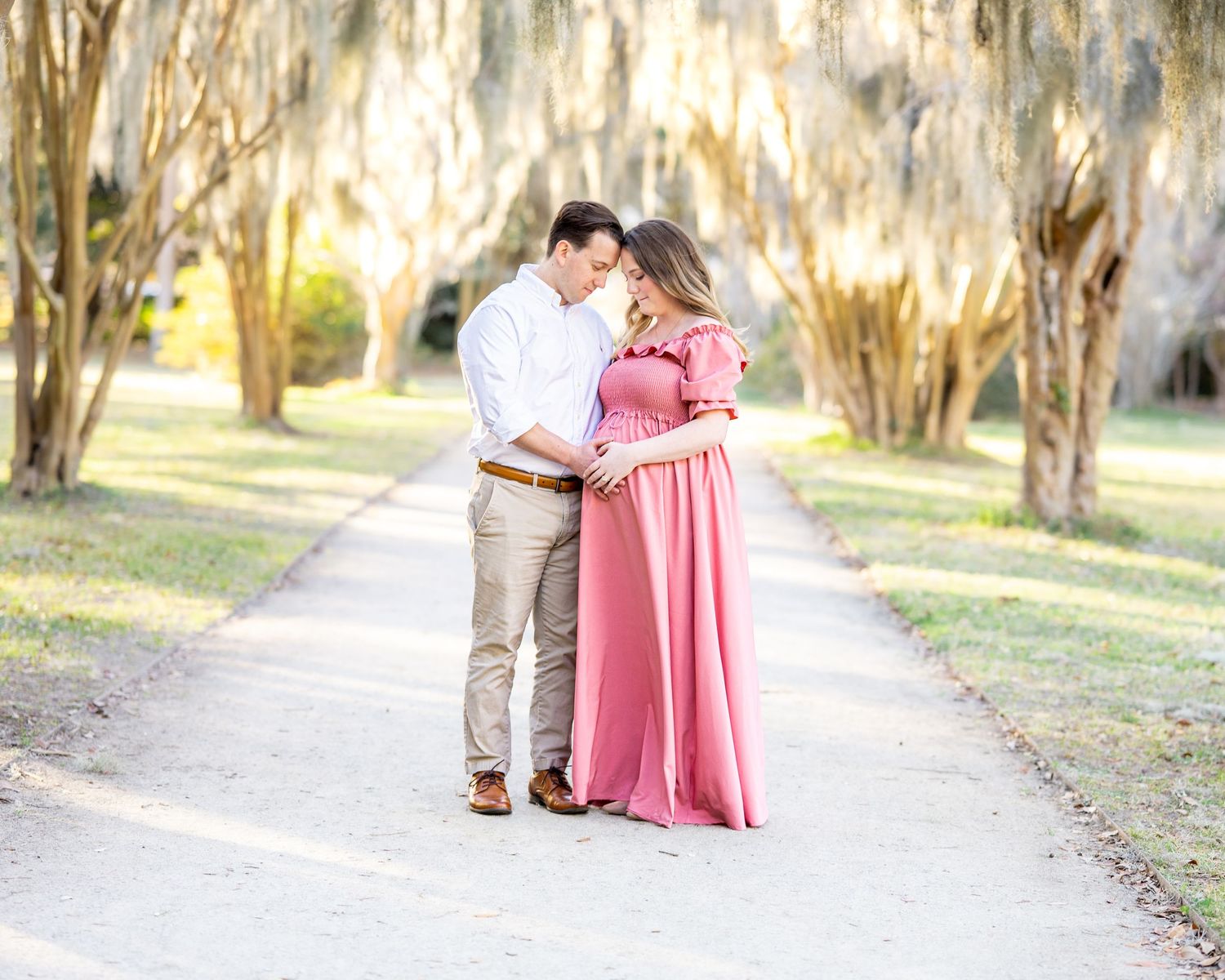 A couple shares a romantic moment on a tree-lined path while sunlight filters through Spanish moss in the background.