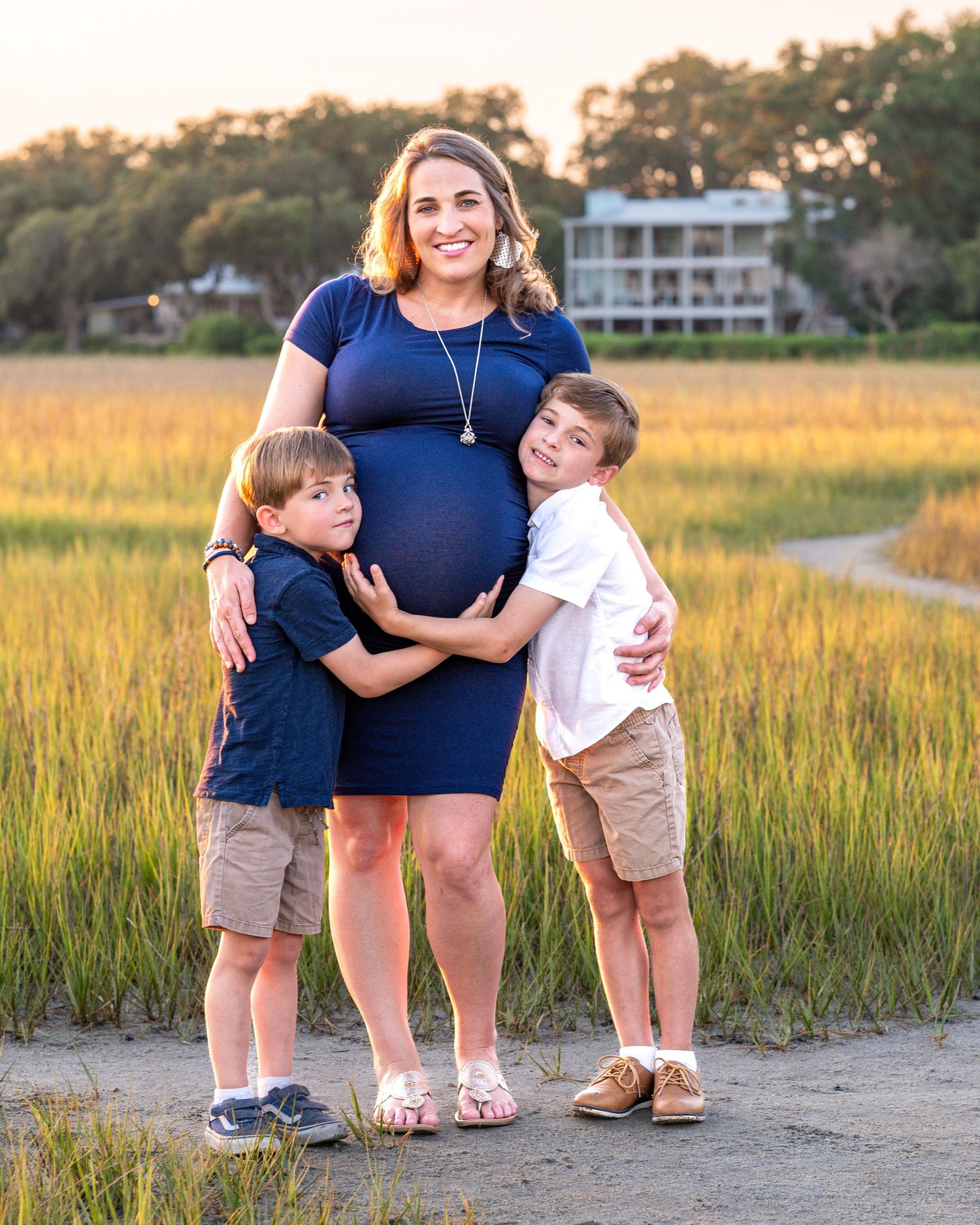 A pregnant woman in a navy dress stands with two young boys on a coastal path at sunset.