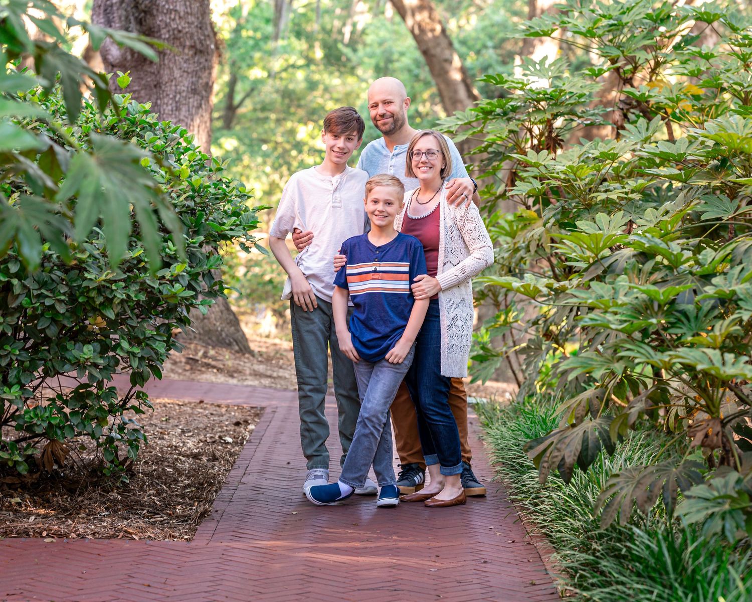 Family of four smiling together on a brick path surrounded by lush green trees and shrubs in a garden setting.