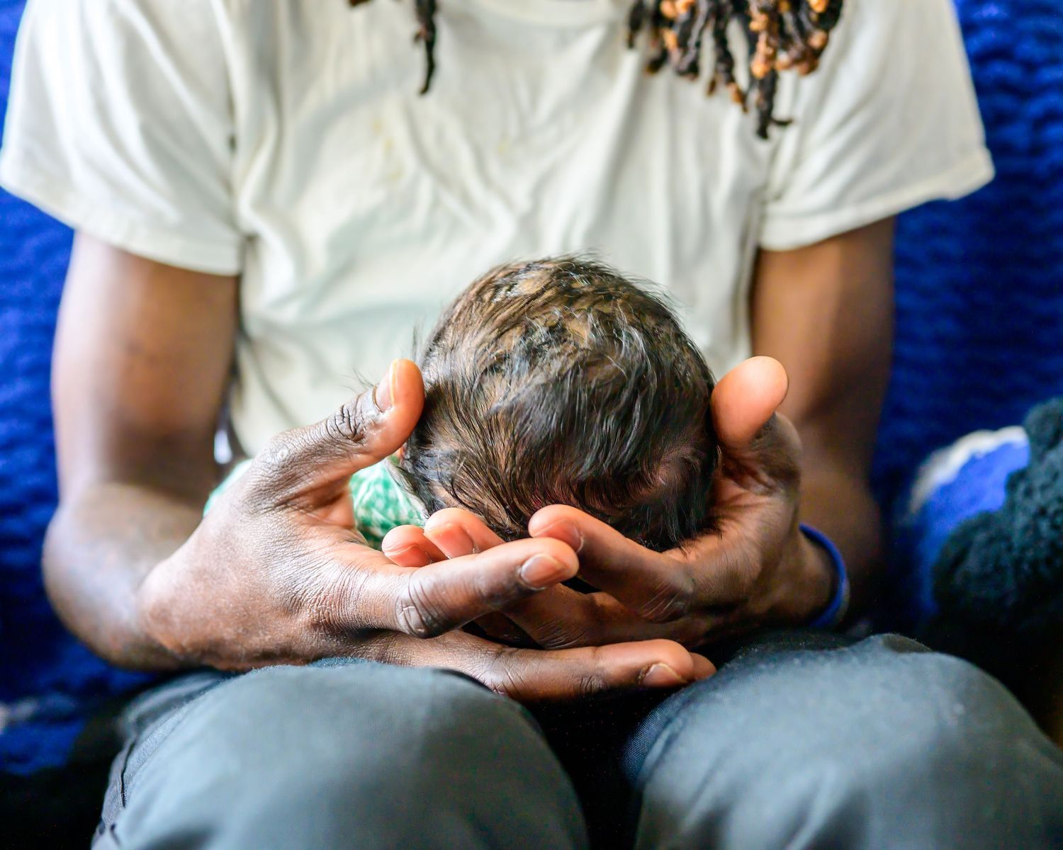 A person tenderly cradling a newborn baby's head in their hands while seated on a blue surface.