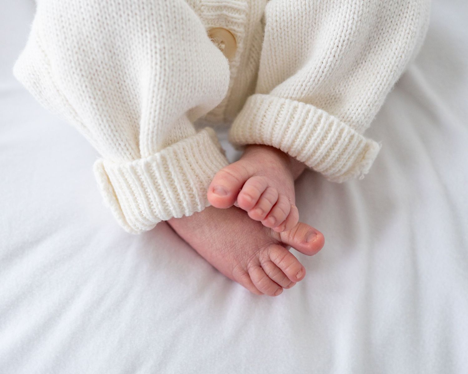 Tiny baby feet wrapped in soft white knitted sweater sleeves against white background.